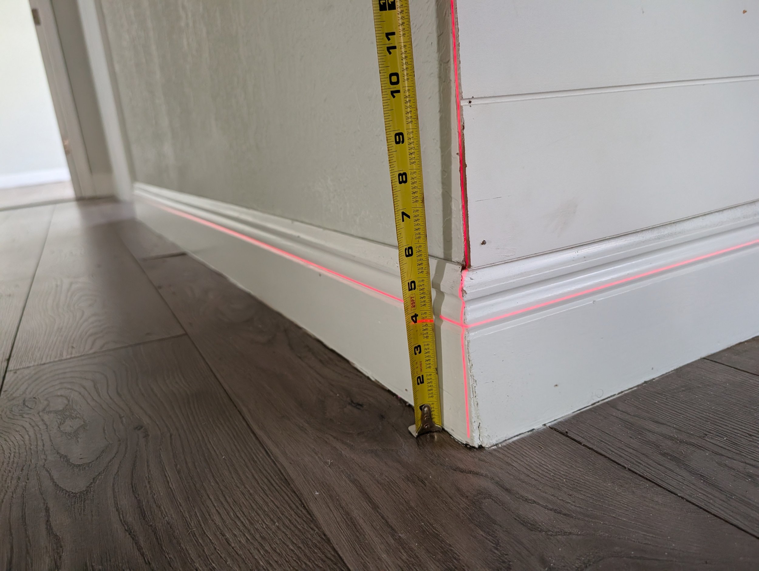A measuring tape with black markings and numbers, positioned vertically against a white wall corner where baseboards meet, with a red laser line marking the measurement line.