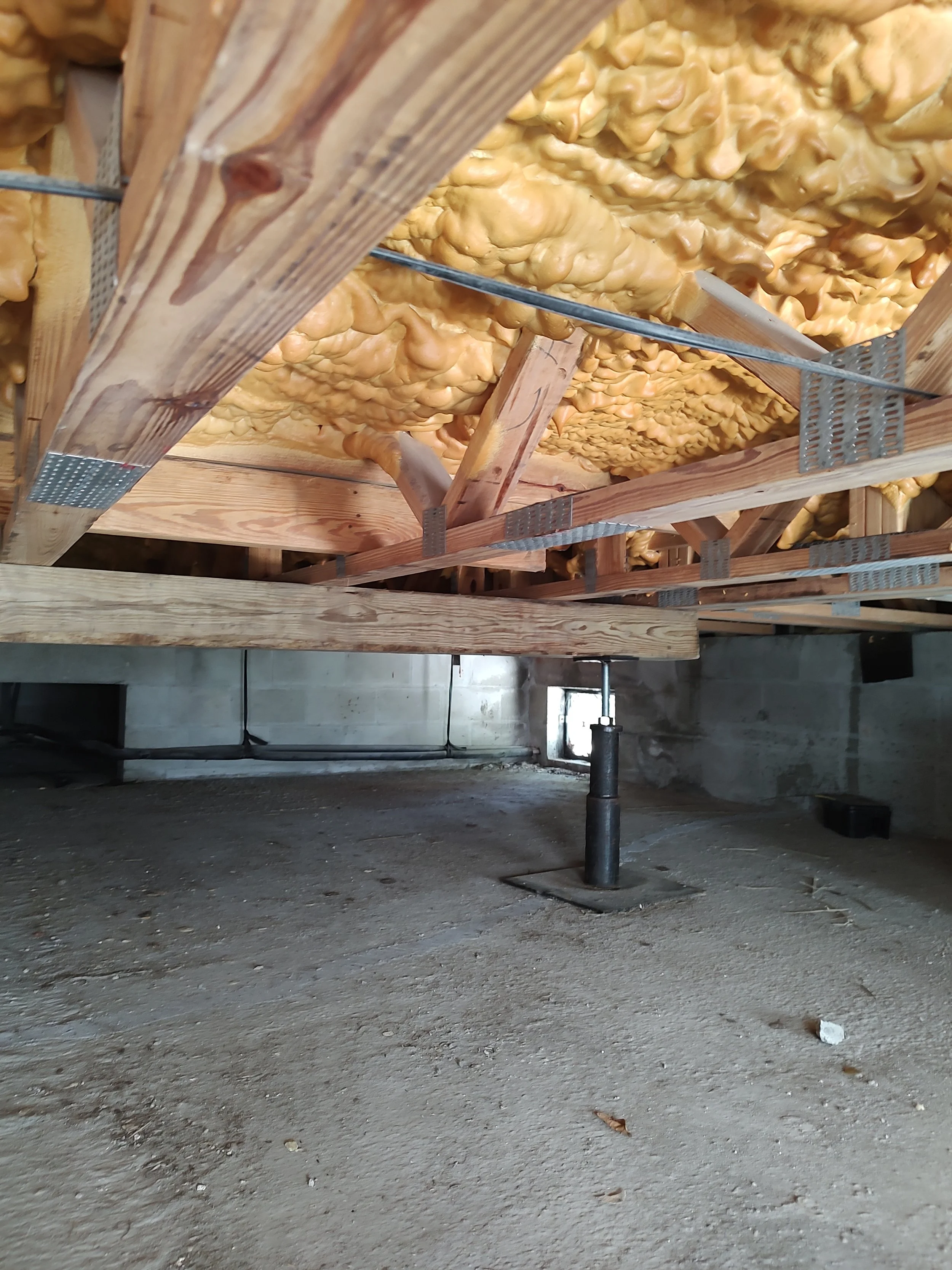 View of a construction site with wooden framing and spray foam insulation on the ceiling, exposed concrete floor, and a small window in the background.