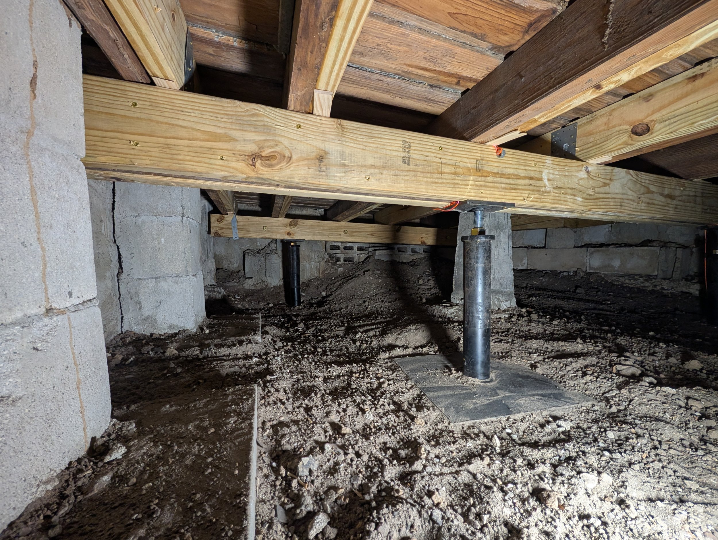 Construction site with wooden beams supported by metal poles and concrete blocks, dirt ground, and an unfinished structure.