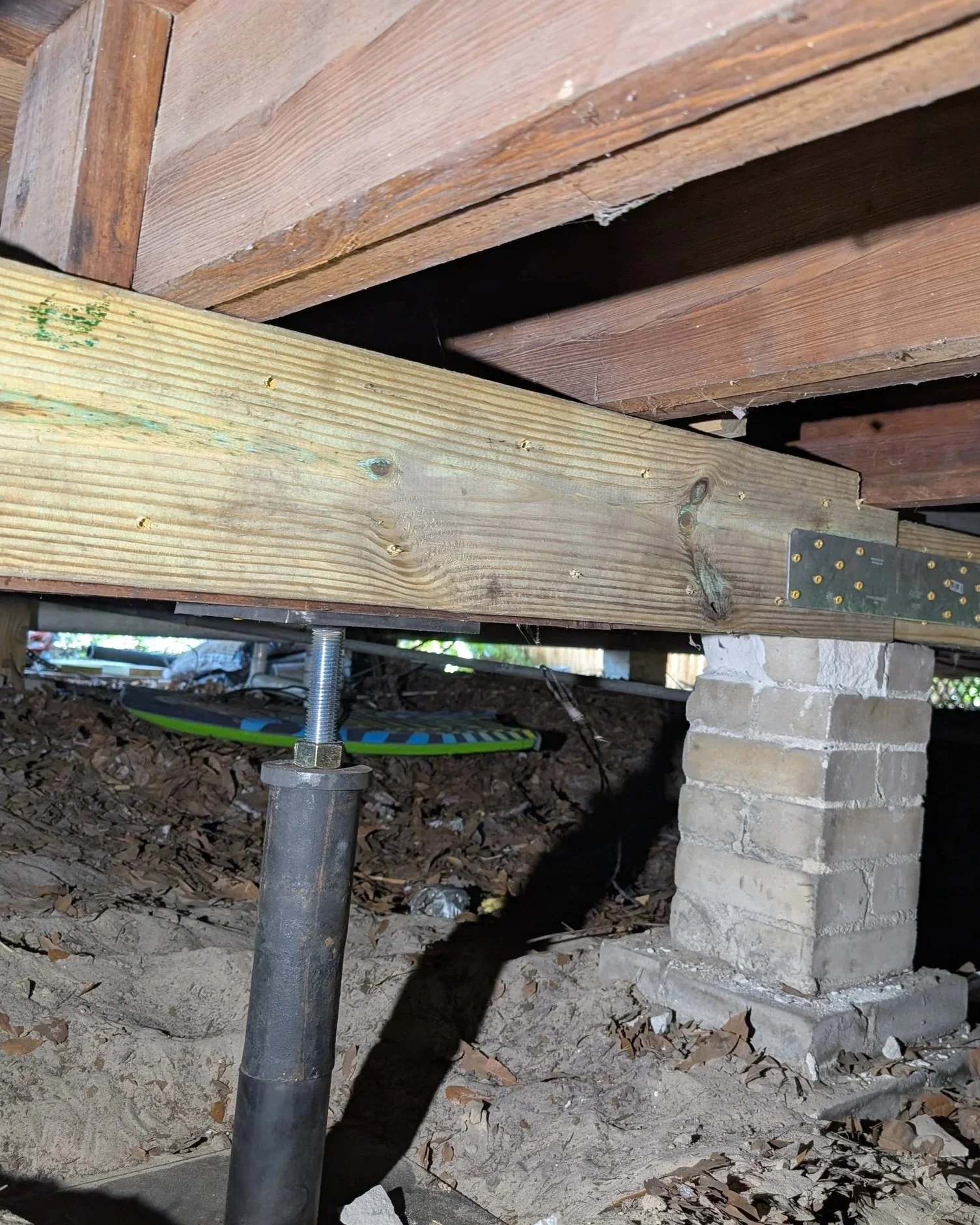 Underpinning of a building or deck, showing a wooden beam supported by a metal post and cinder block foundation with leaves scattered on the ground.