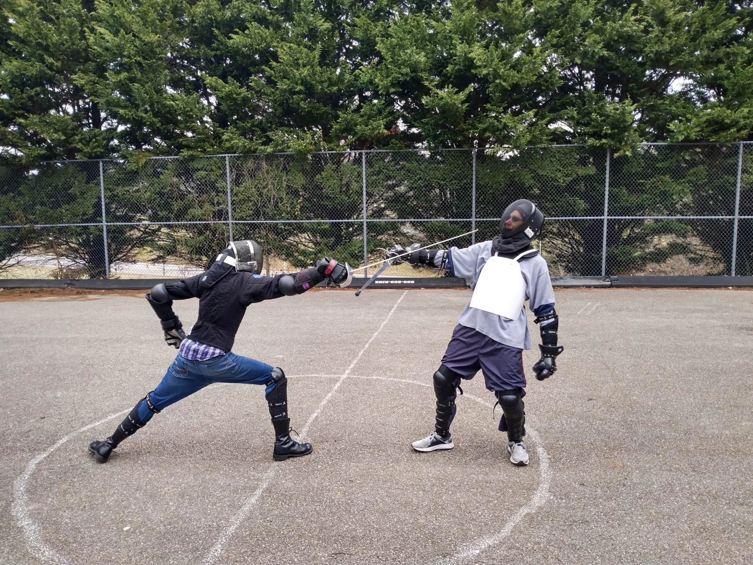 Two individuals engaging in a fencing duel outdoors, wearing protective gear, including helmets and gloves, with green trees and a chain-link fence in the background.