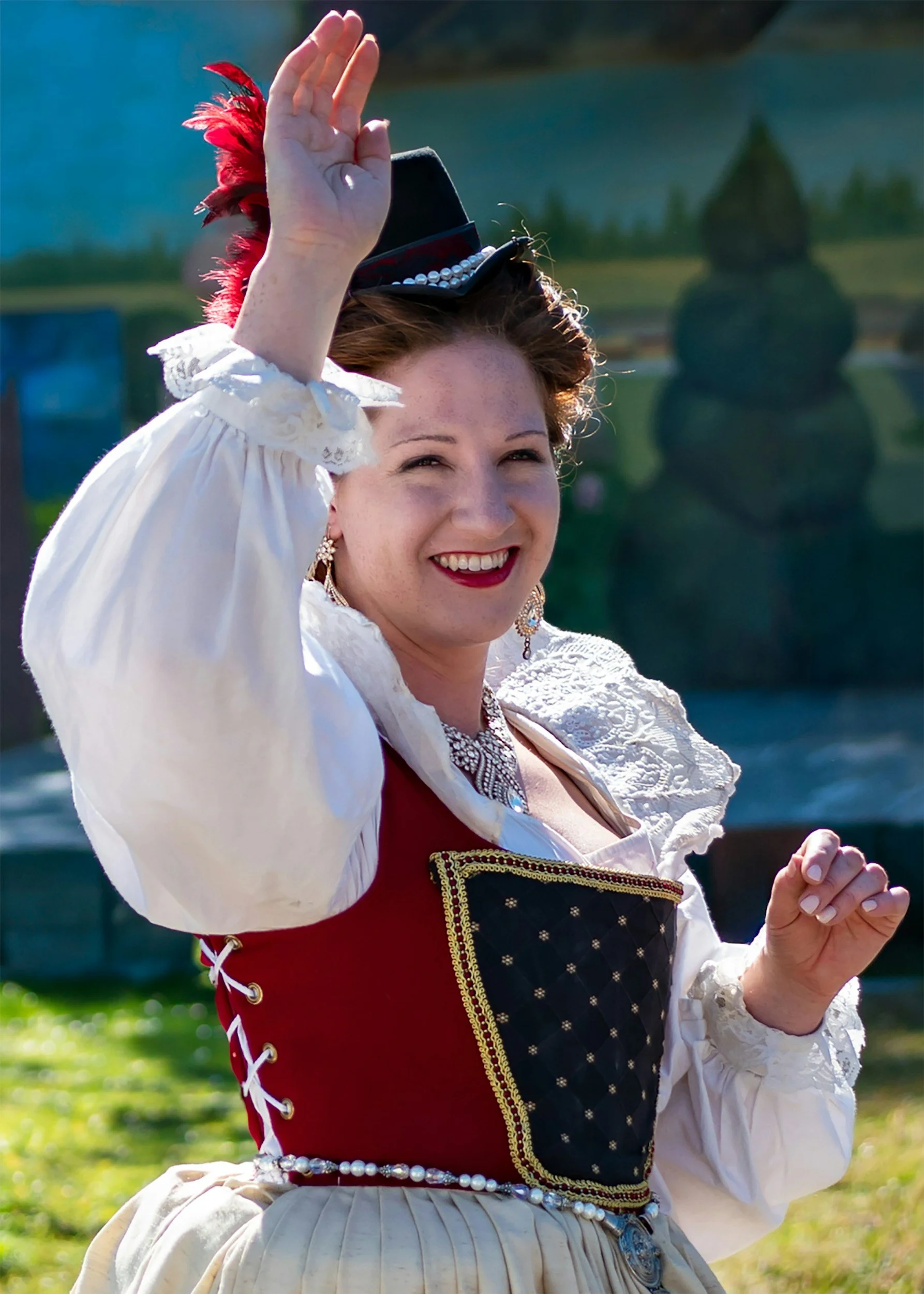 A woman dressed in traditional European folk costume, smiling and waving, wearing a black hat with a red feather, a white blouse with lace, and a decorative vest with gold trim, outdoors with a body of water and greenery in the background.