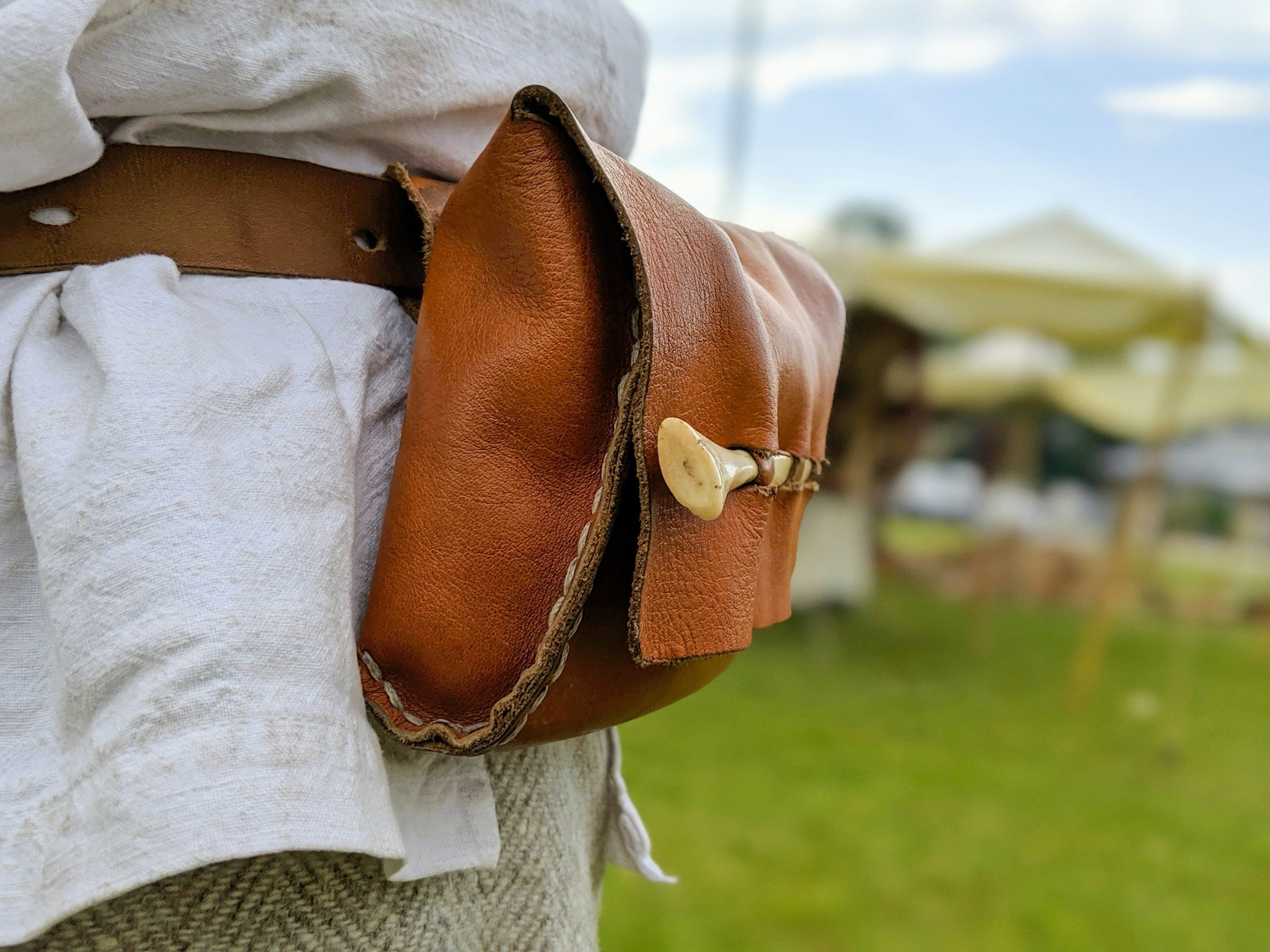 Close-up of a person wearing a white shirt, tan belt, and carrying a brown leather bag with a horn-shaped clasp, holding it against their side. The background is blurred with a cloudy sky and some greenery.