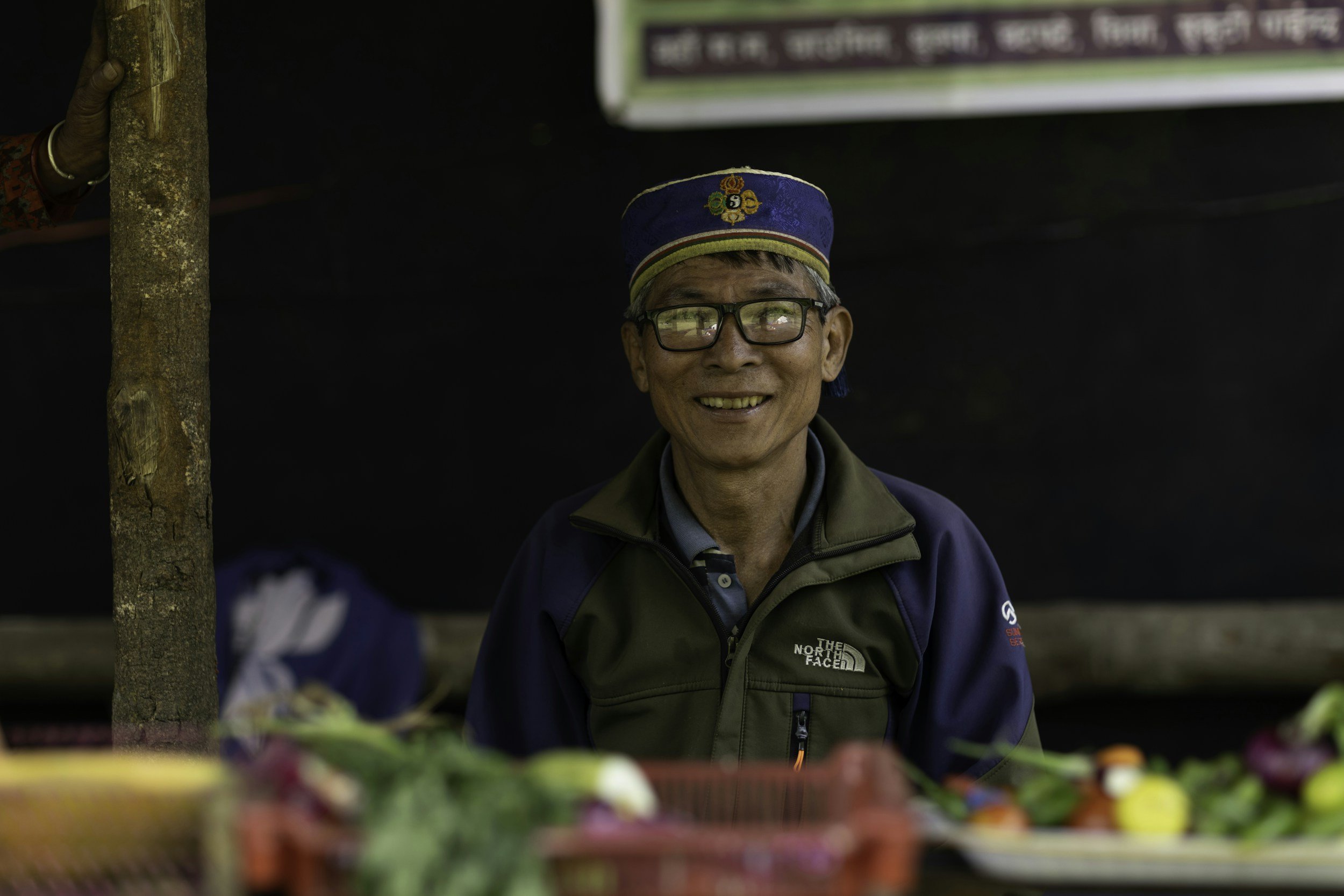 A smiling elderly man wearing glasses, a blue and black North Face jacket, and a traditional Nepalese hat, standing behind a table with colorful fruits or flowers at an outdoor market.