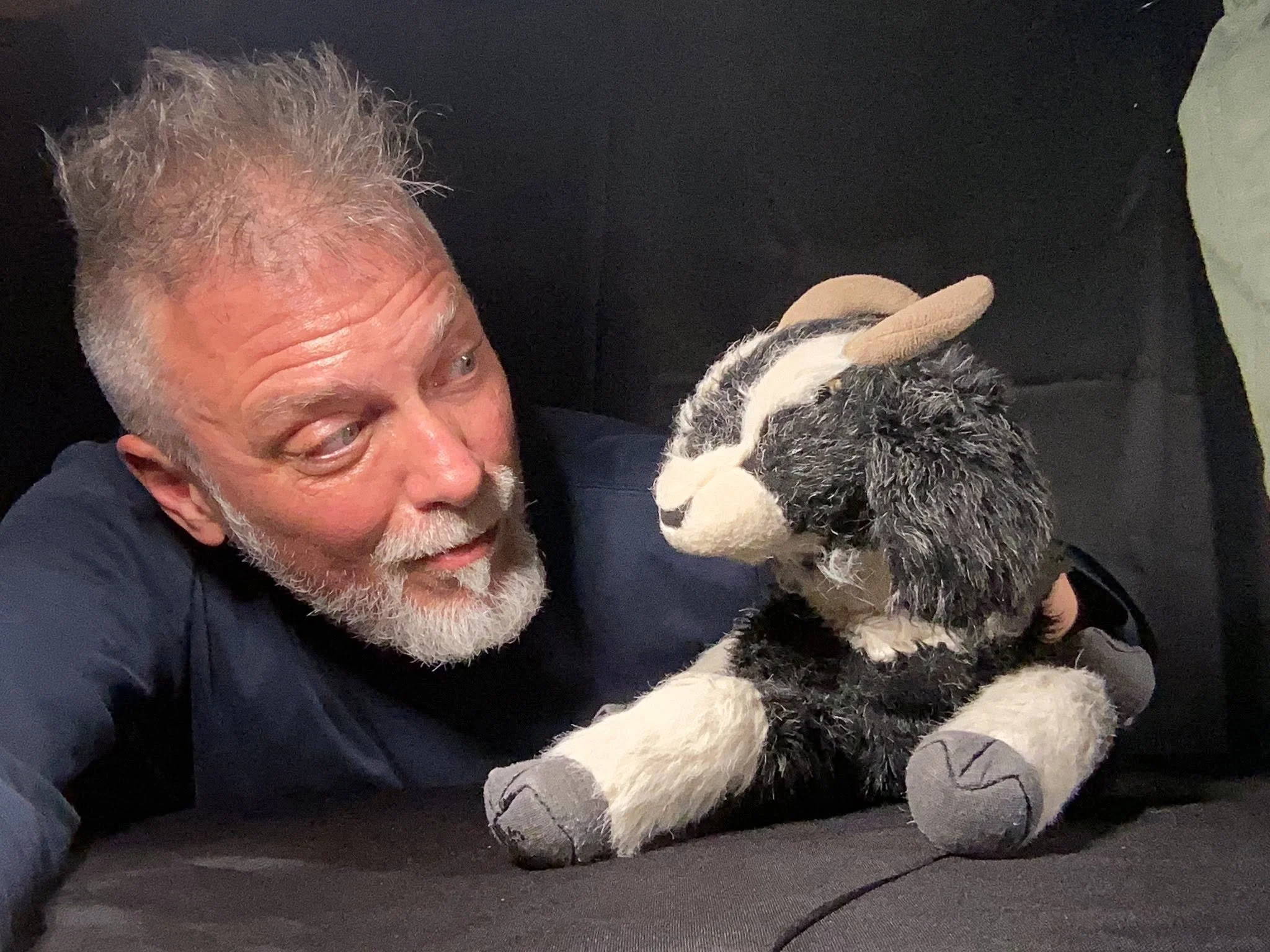 A man with white hair and a beard looking at a puppy. The puppy has a black and white fur pattern and is wearing small socks on its paws. They are on a dark surface with a dark background.