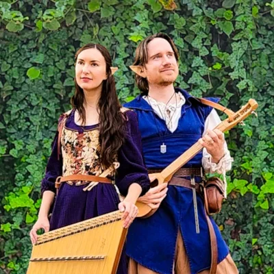 A man and woman dressed in medieval-style costumes stand outdoors against a leafy green background, holding musical instruments.