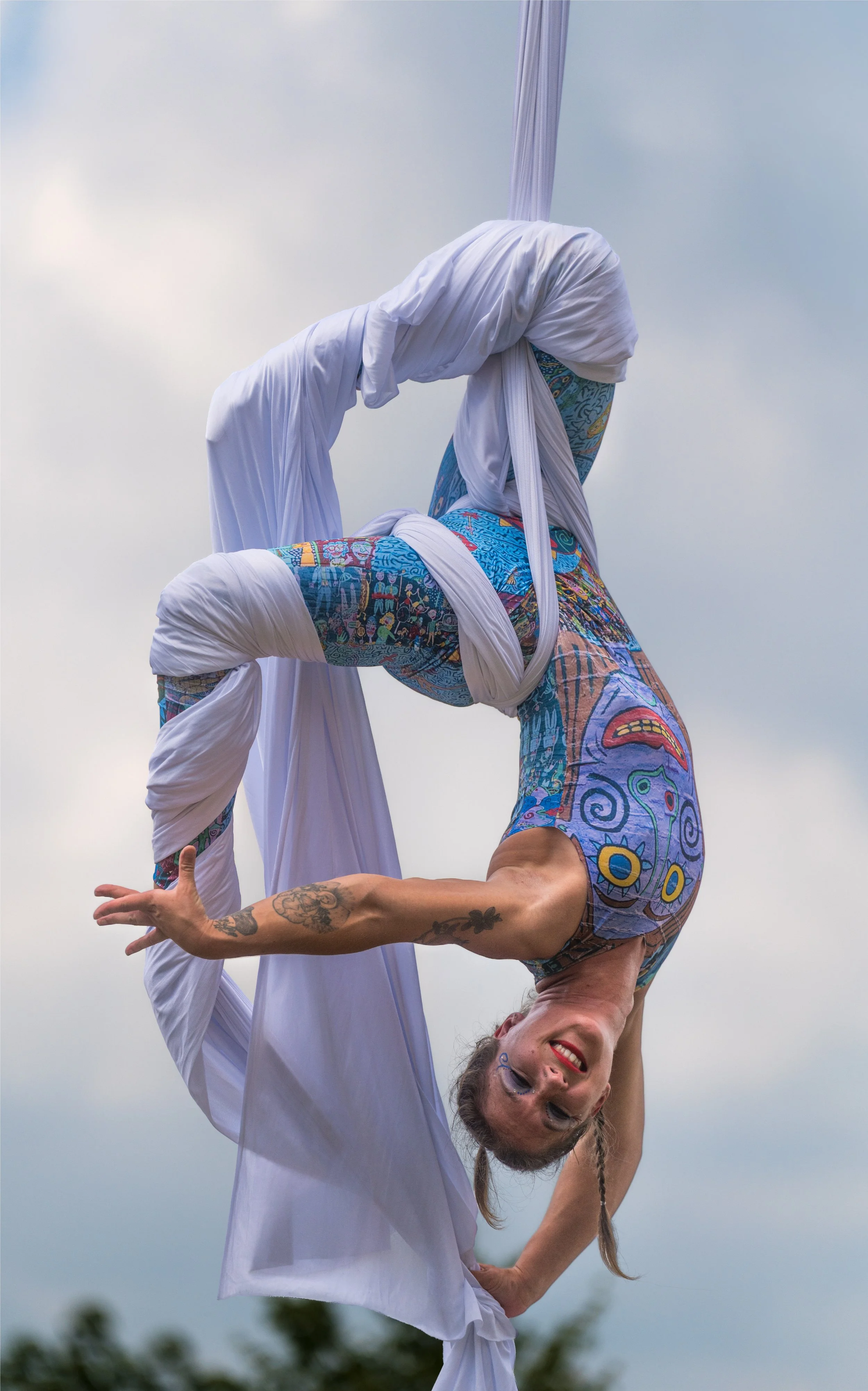Two women practicing acro yoga outdoors, one in a white outfit balancing upside down while holding the other with tattoos in colorful, patterned clothing, under a cloudy sky.