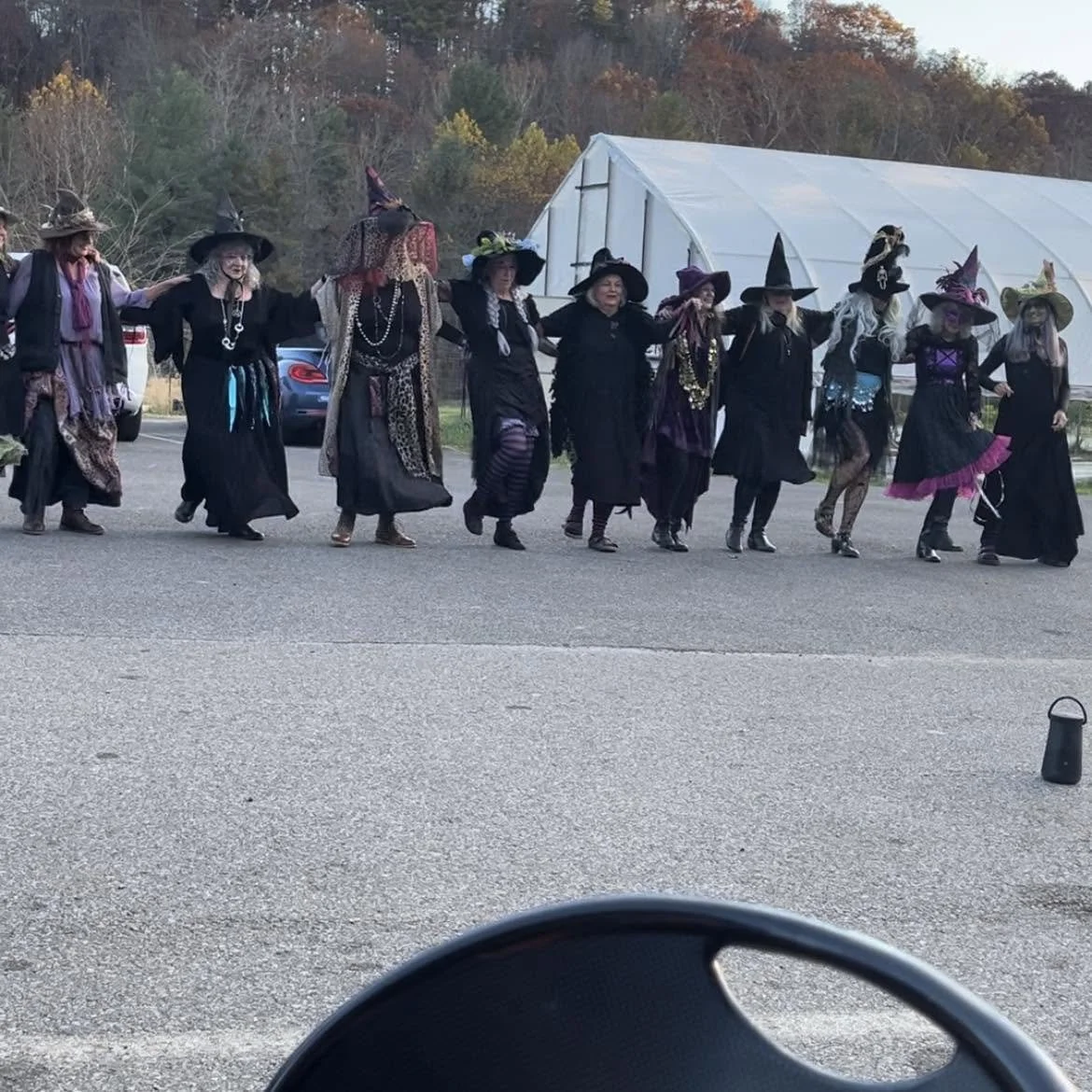 A group of people dressed as witches holding hands in a line, participating in a Halloween or costume event outdoors, with a tent and trees in the background.