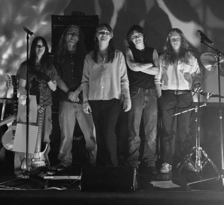Five women standing on a stage with musical instruments and microphones, performing or preparing to perform in a dimly lit setting.