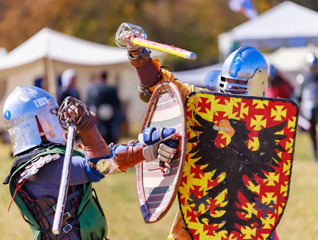 Two individuals in medieval armor engaging in a mock sword fight at an outdoor event, with tents and people in the background.