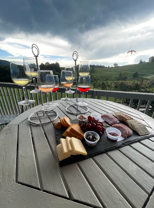 A table with wine glasses, cheese, grapes, and charcuterie with a scenic outdoor view of green hills and a cloudy sky.