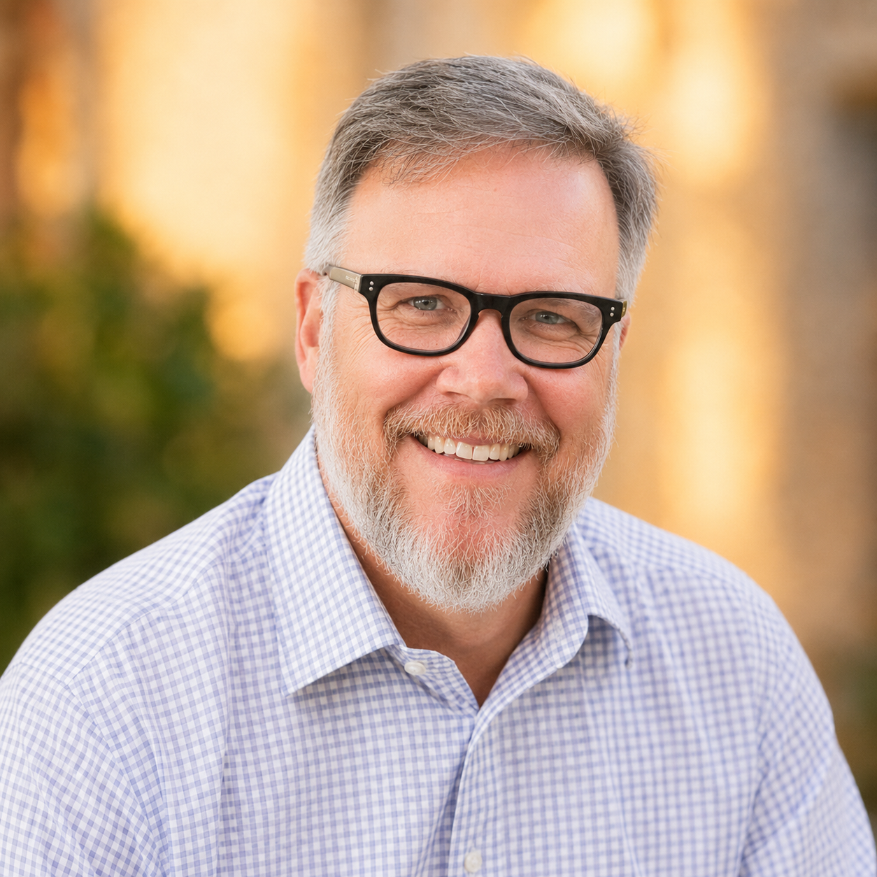 Portrait of a smiling middle-aged man with gray hair, beard, and glasses, wearing a light blue checkered shirt, outdoors with blurred warm-colored background.