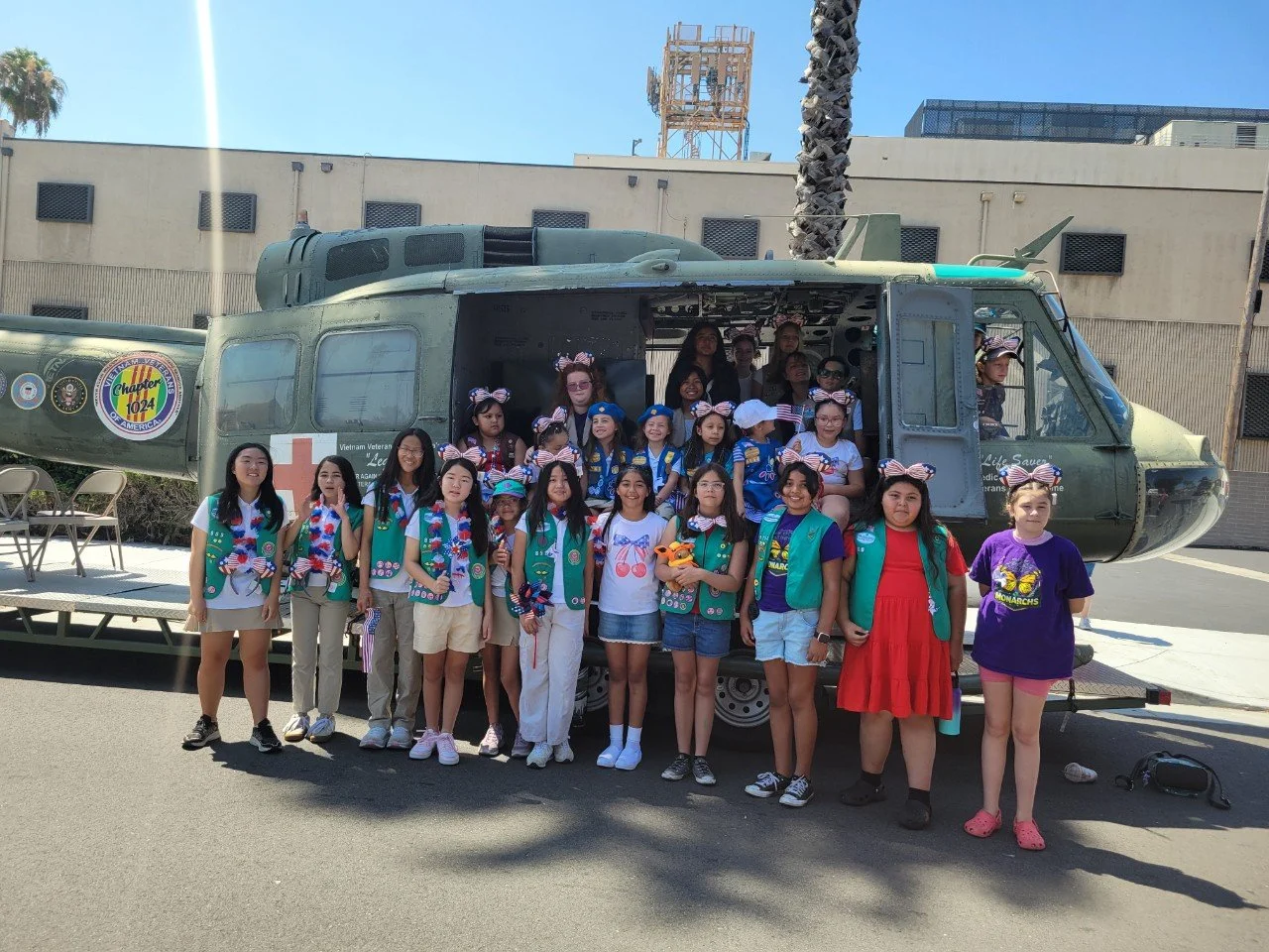Group of children standing in front of a military helicopter, some wearing Girl Scout uniforms and bows, outdoors on a sunny day.