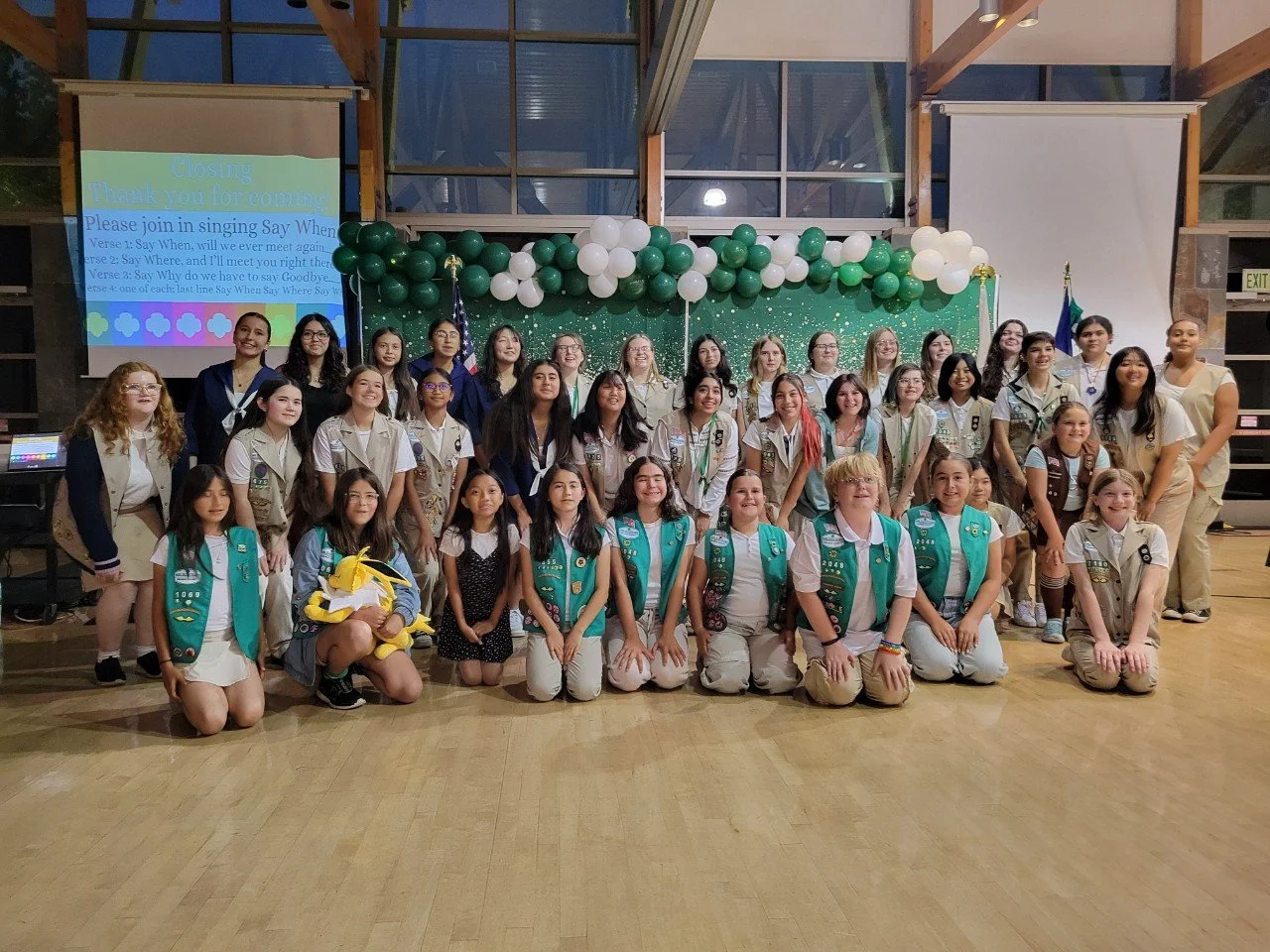 A group of girls dressed in Girl Scout uniforms and casual clothing posing for a photo on a wooden floor in front of a decorated stage with green and white balloons, a green backdrop, and a projector screen displaying song lyrics.