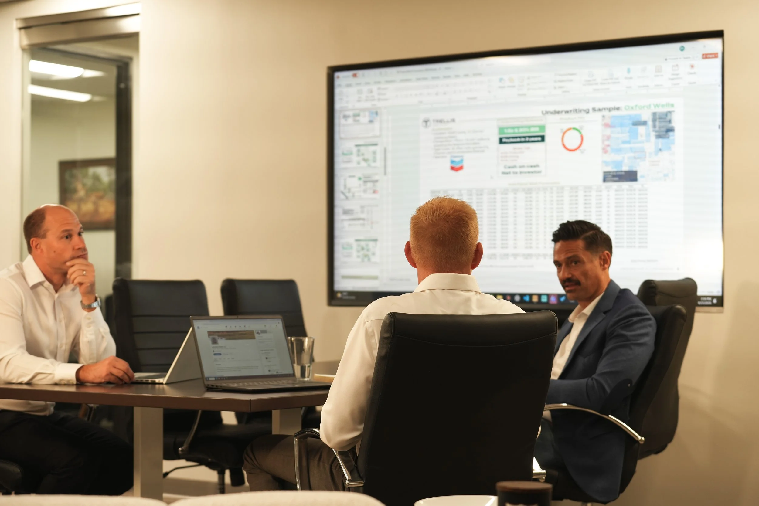 Four men in a business meeting or discussion around a conference table, with laptops and a large monitor displaying data and charts in the background.
