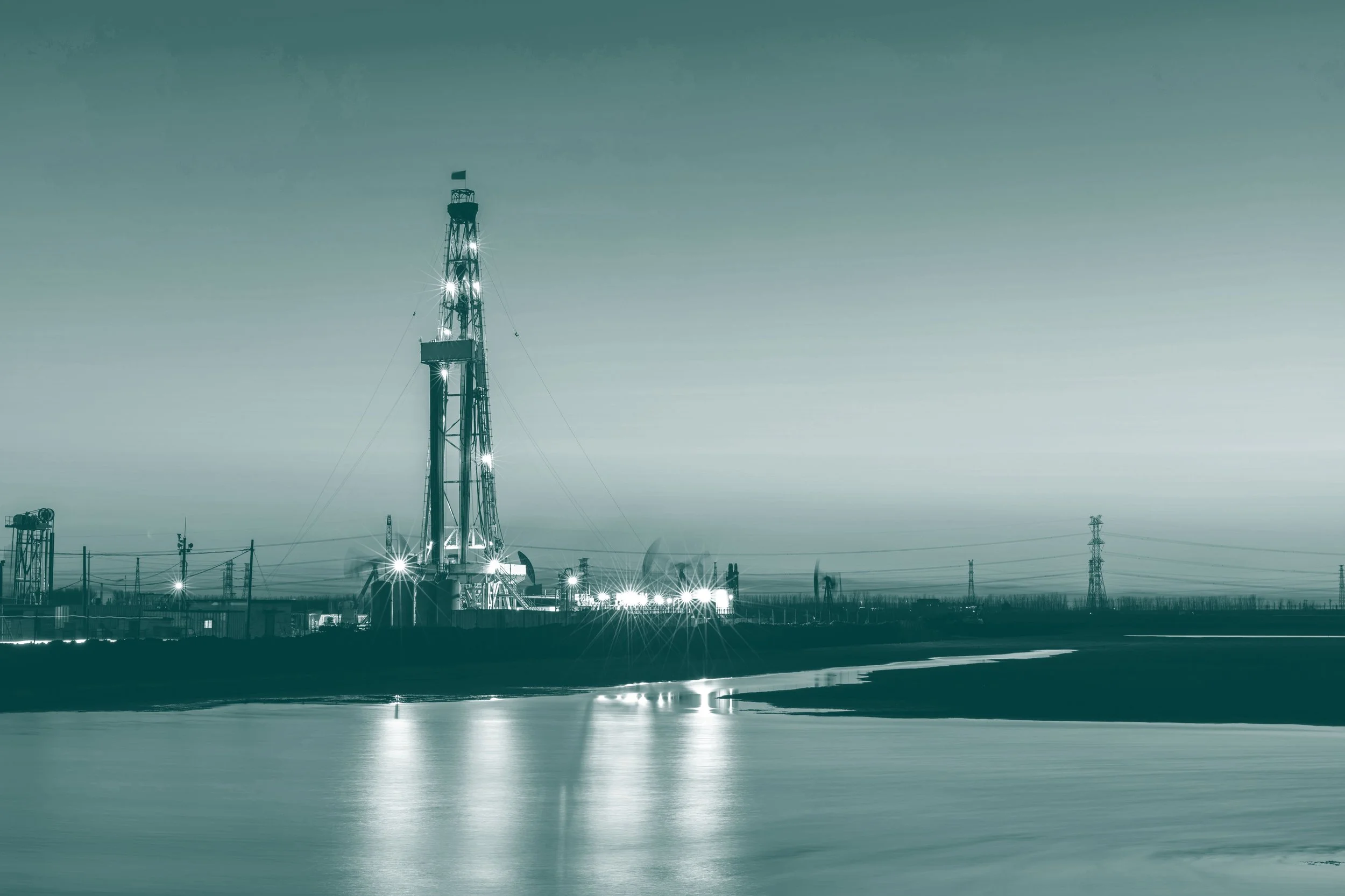 An oil and gas operation site, surrounded by power lines and infrastructure, reflected on a calm body of water, with an overcast sky in the background.