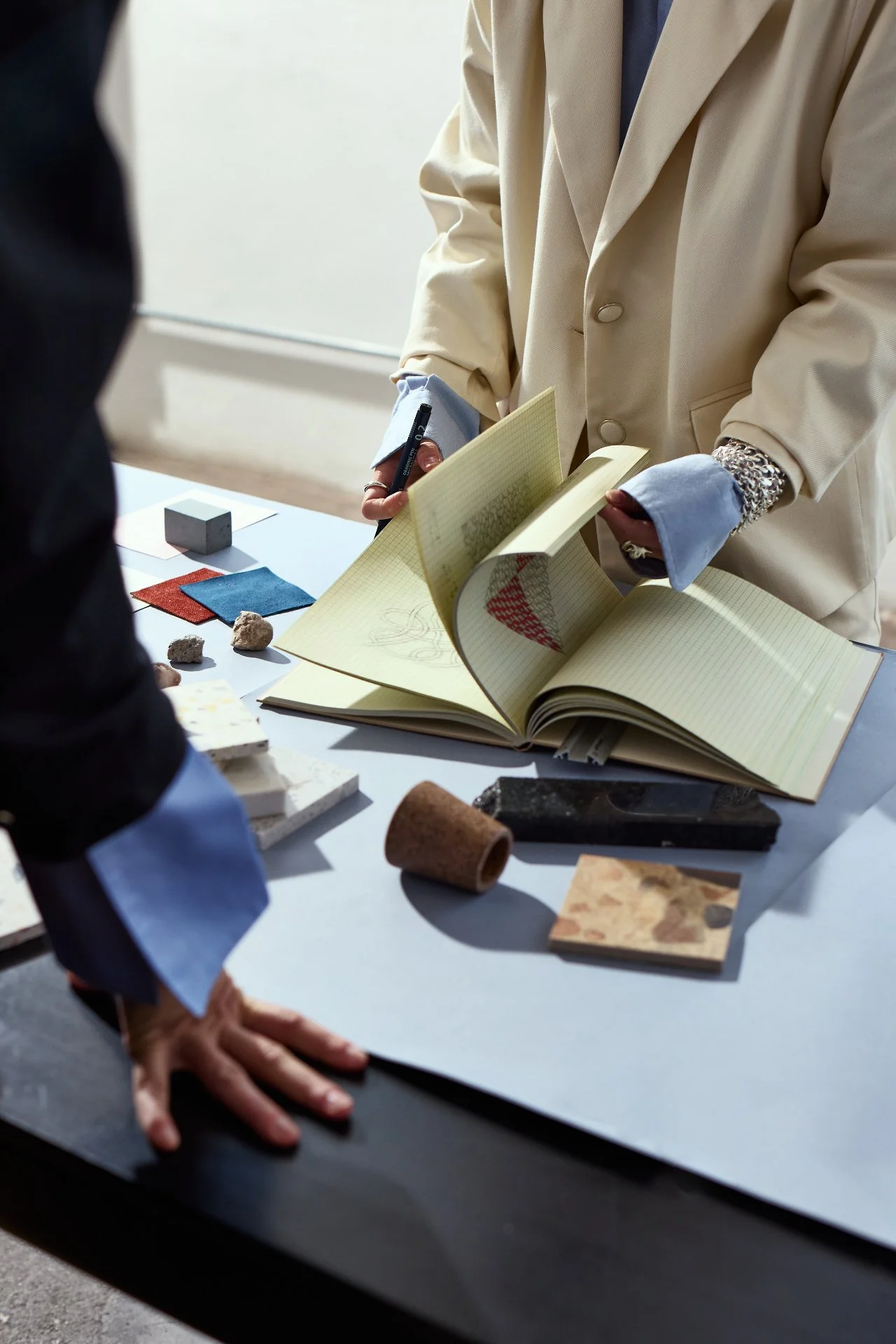 Close-up of a person wearing a beige blazer holding an open notebook and a pen, with a table nearby displaying various fabric samples and small objects, in a well-lit room.
