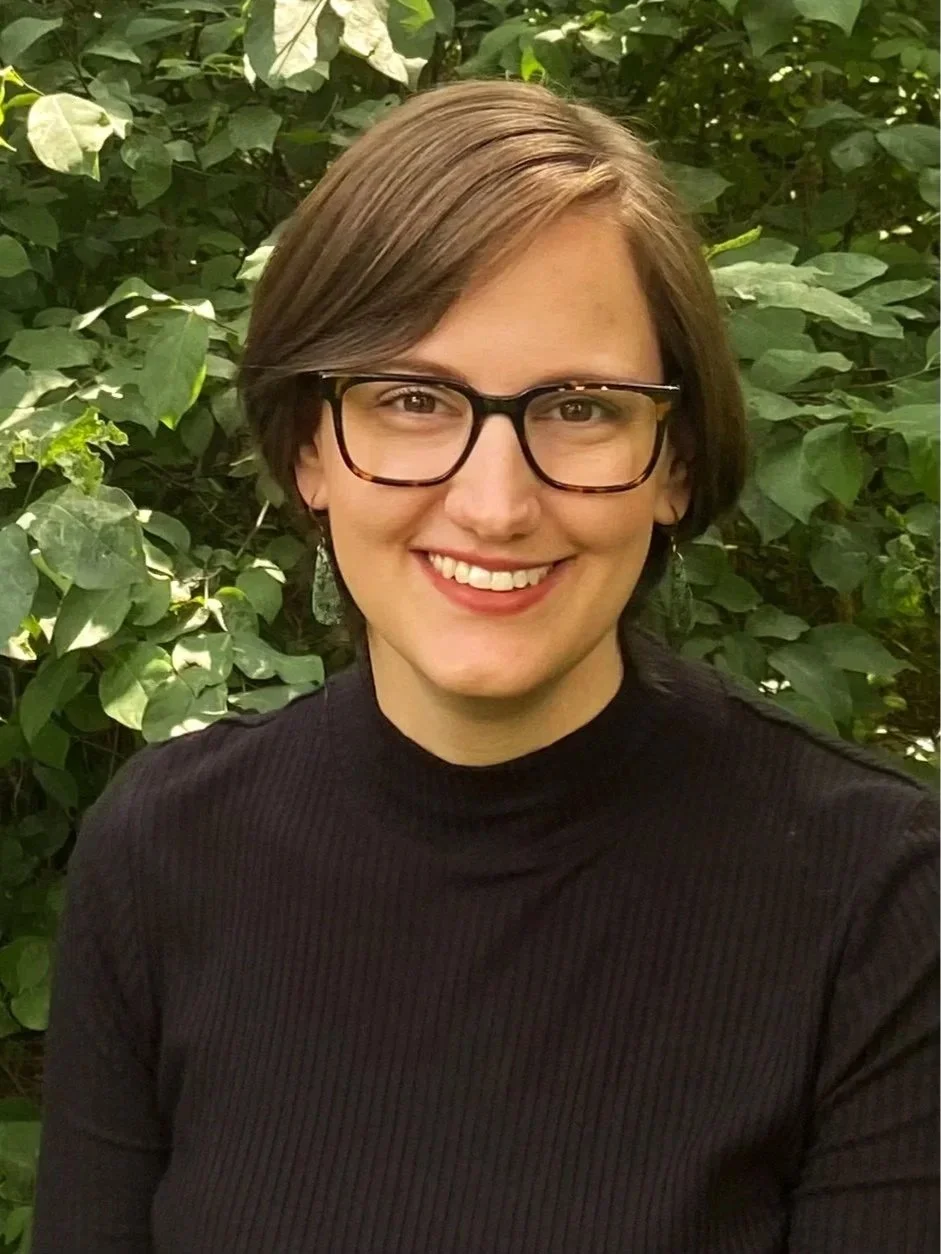 A woman with short brown hair, glasses, and earrings smiling in front of green leafy bushes, wearing a black ribbed top.