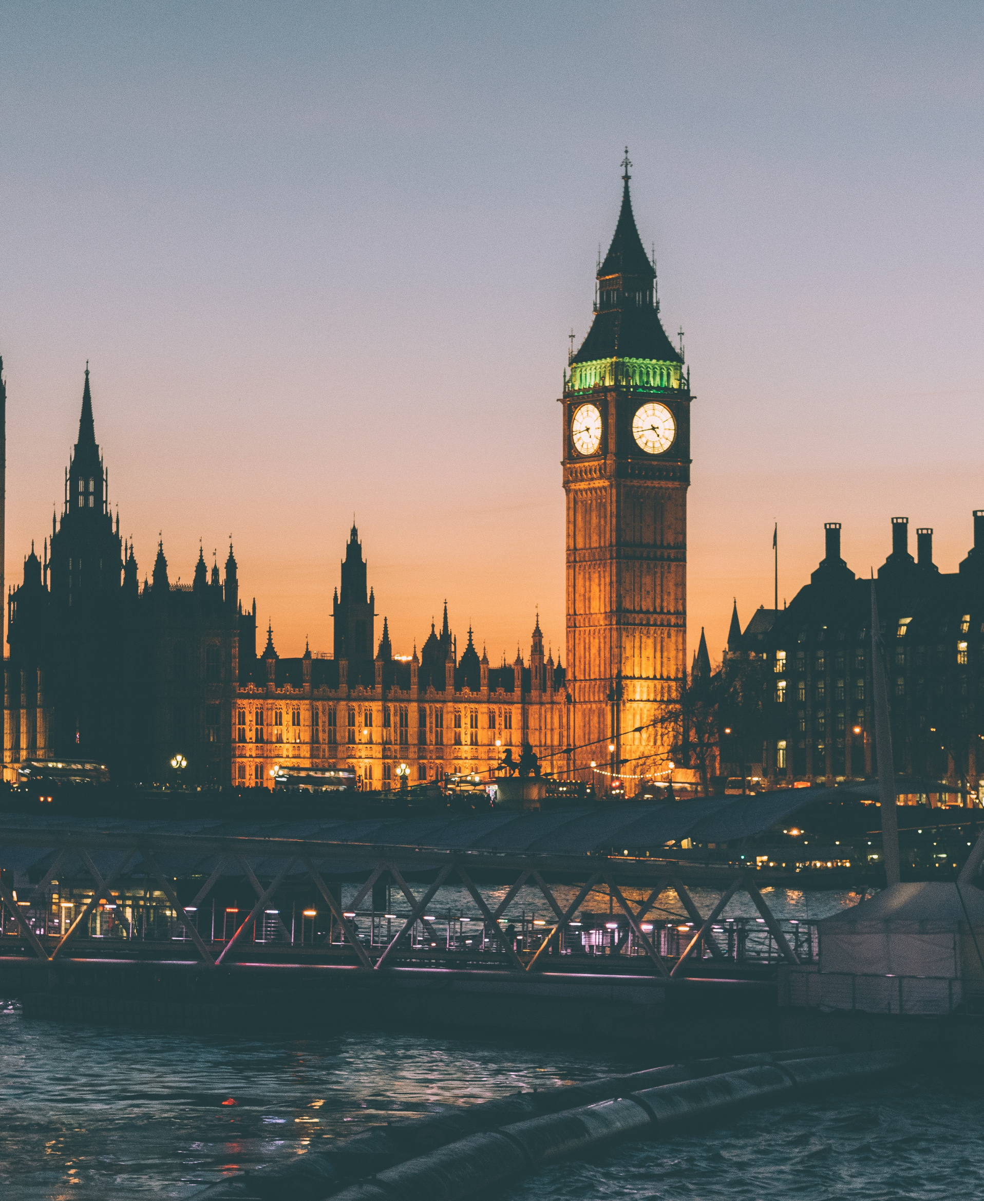 Cityscape featuring Big Ben clock tower illuminated at dusk, with a bridge over water in the foreground.