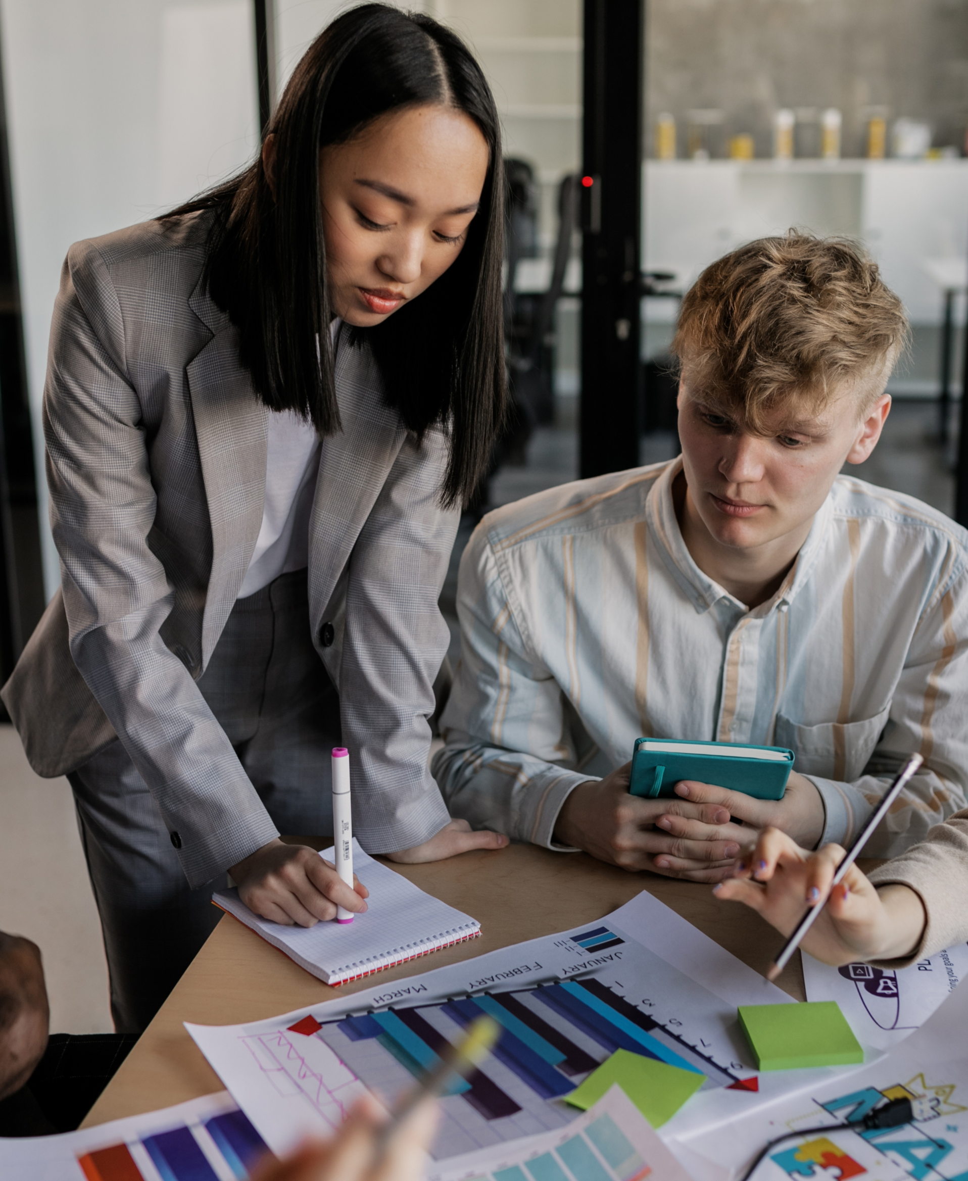 Two people working together at a table covered with colorful charts, graphs, and notes, with one person holding a tablet and the other writing on a notepad.