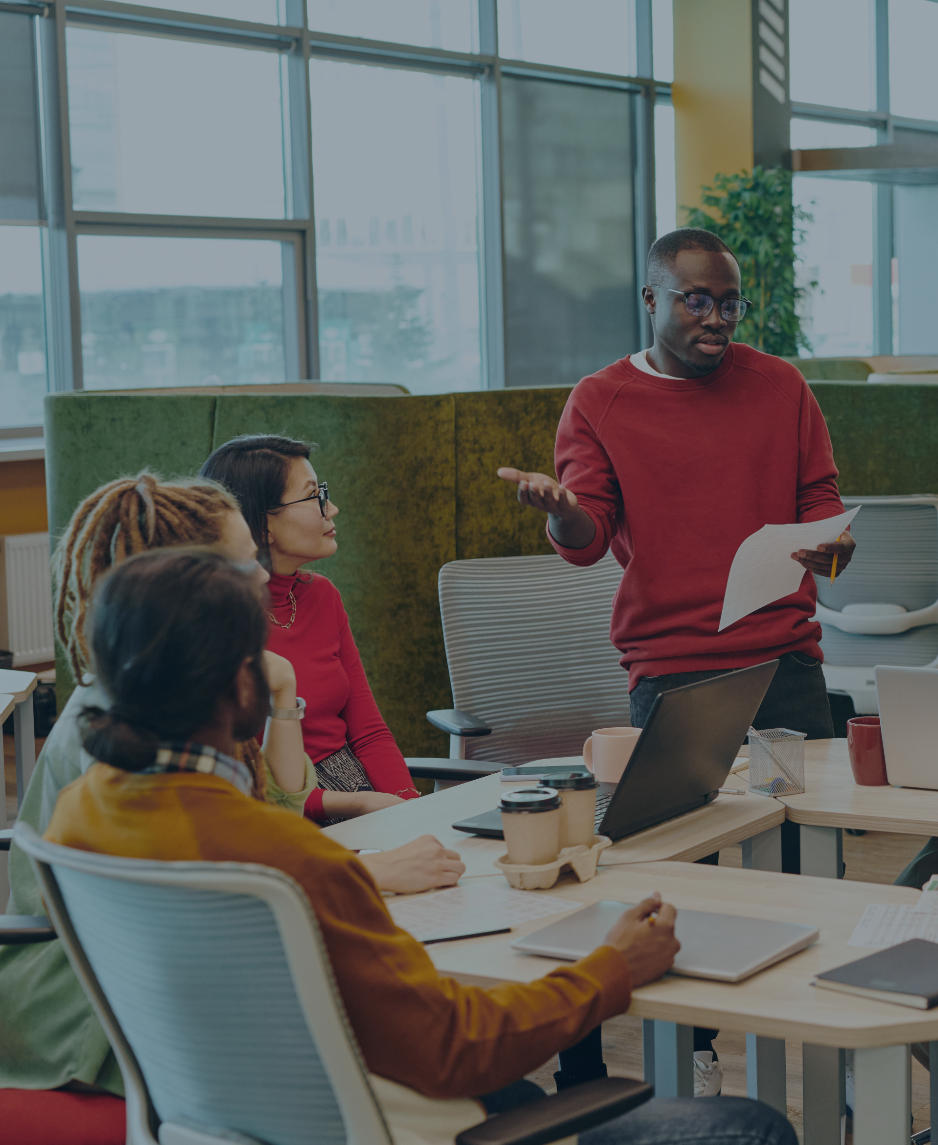 A group of diverse people in a modern office meeting room, with someone standing and speaking, while others listen, take notes, or work on laptops at a large table.