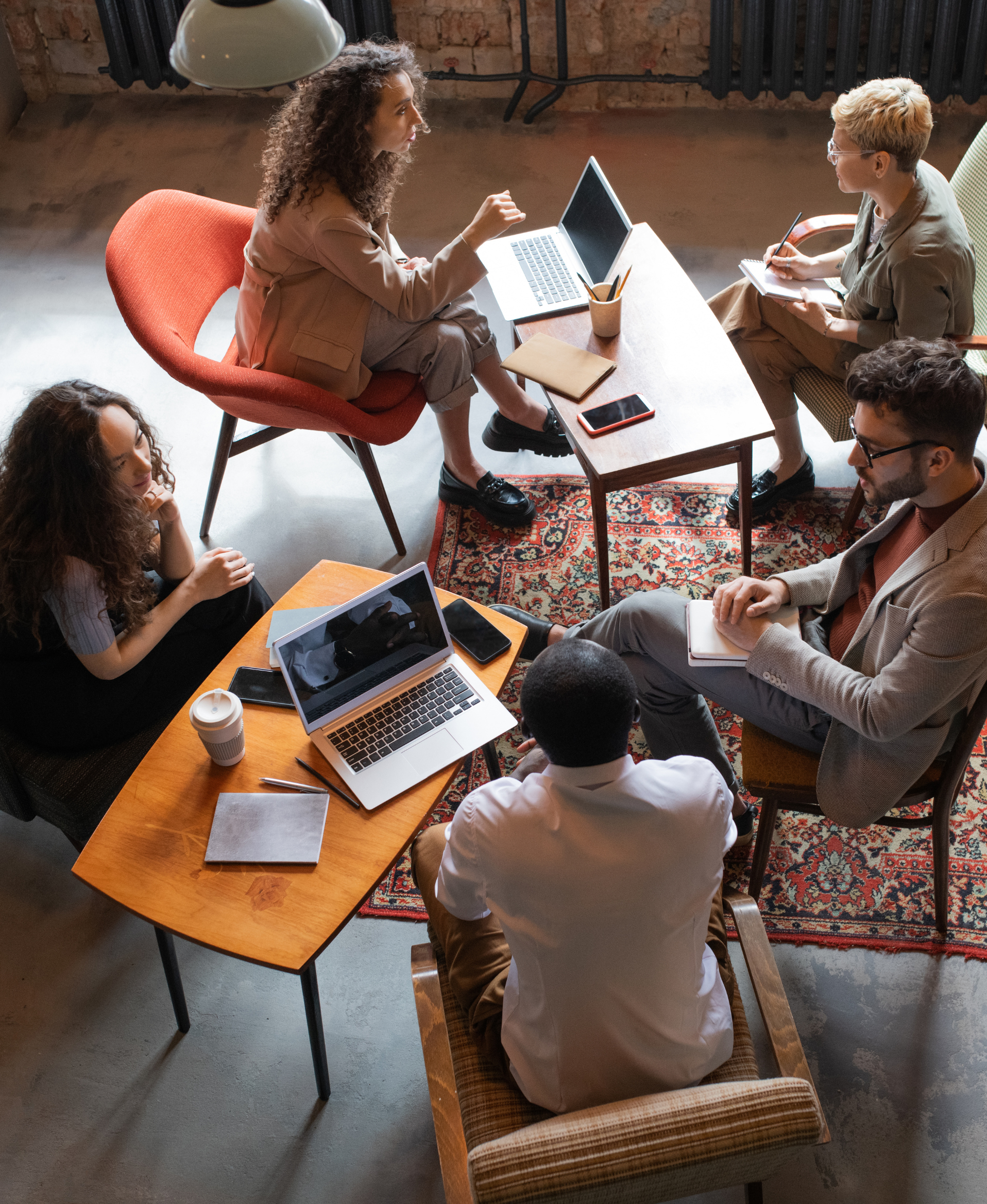 A group of people in a meeting or discussion in a room with brick walls and a large window. They are sitting around a wooden table with laptops, notebooks, and beverages.