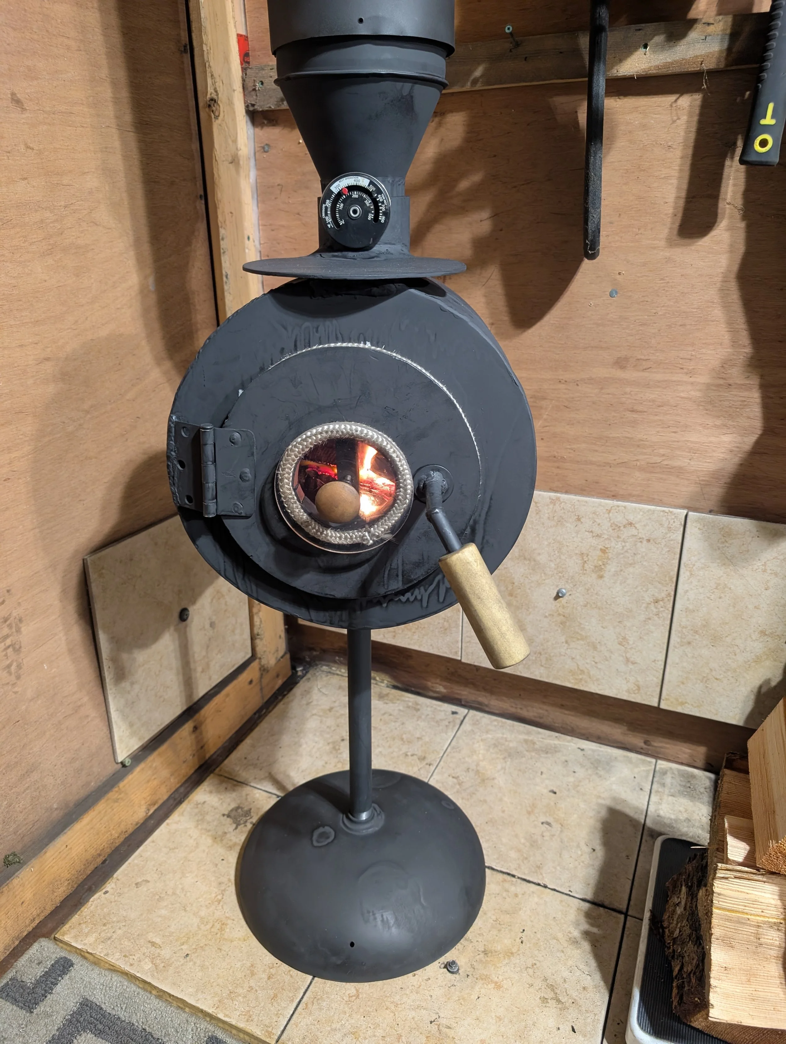 A black, round, wood-burning stove with a metal pipe and a thermometer attached on top, placed outdoors on a tile and wood surface, surrounded by wood and a black stand.
