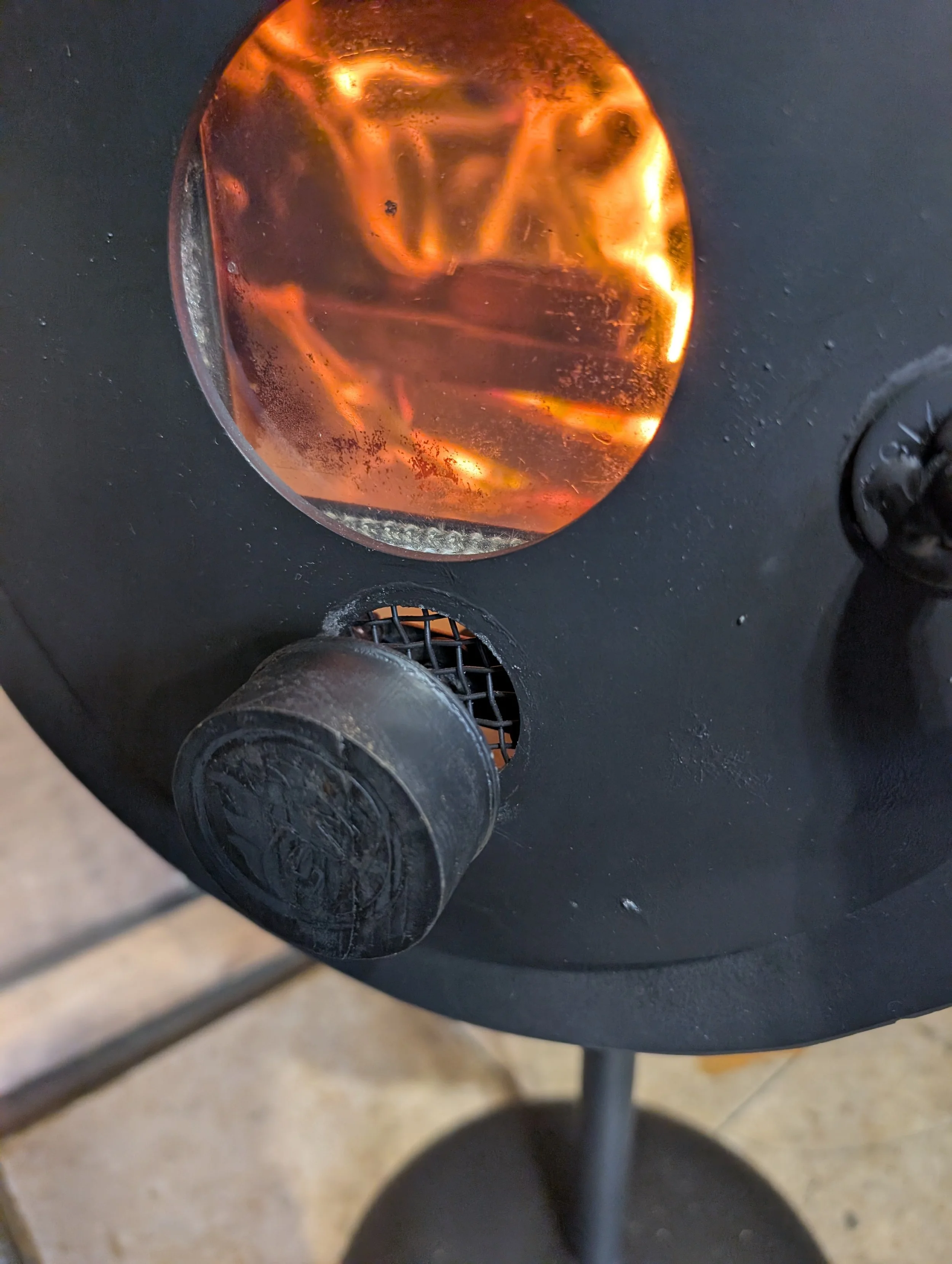 A close-up view of a black tabletop with a metallic object and a glass containing a dark liquid with ice. The photo is taken from an angled perspective showing the edge of the table and part of the floor.