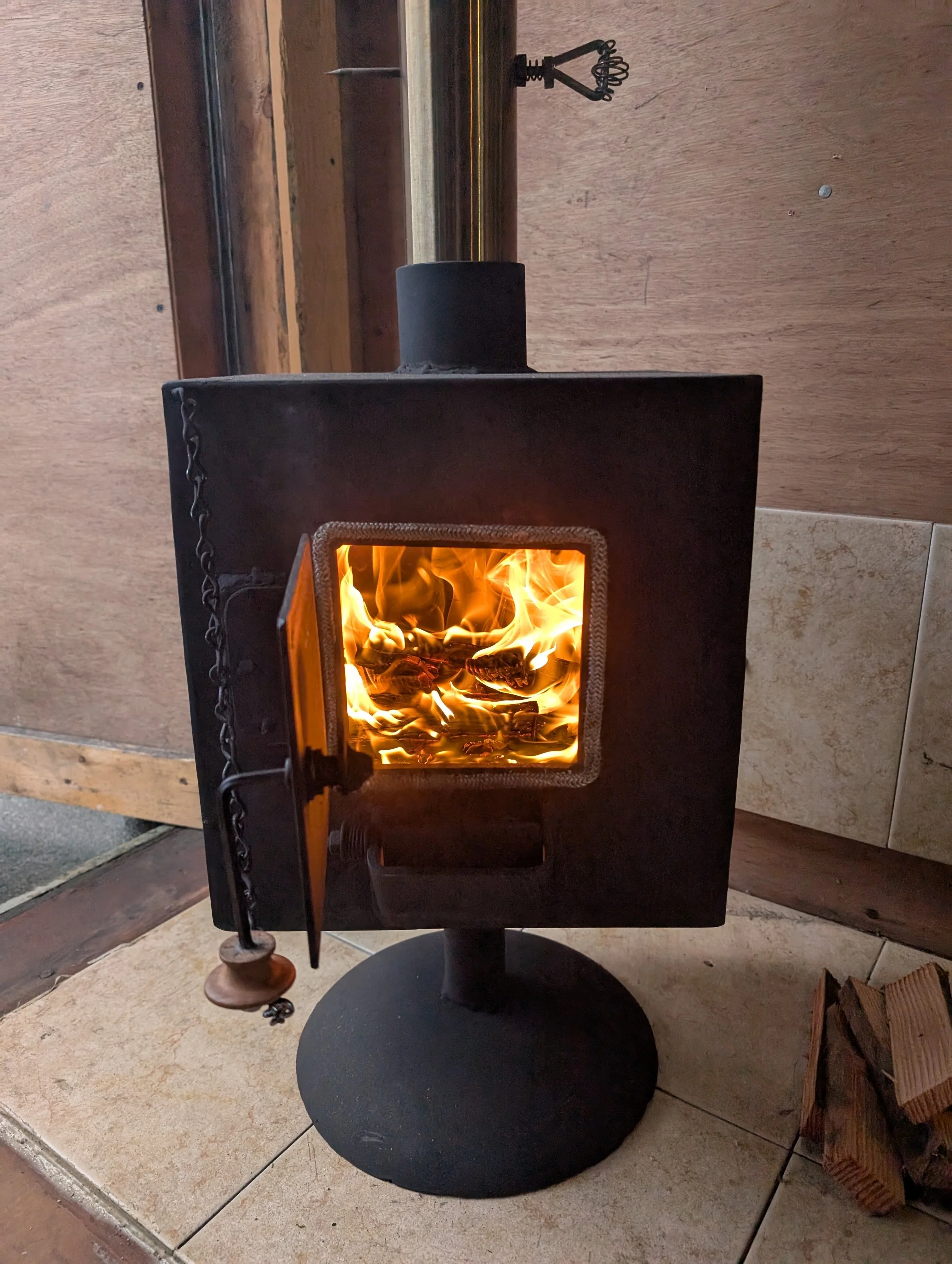 A black cast iron wood-burning stove with a visible fire inside, positioned on a beige tiled floor. The stove has a small chain hanging on the left side and is placed against a wooden wall background.