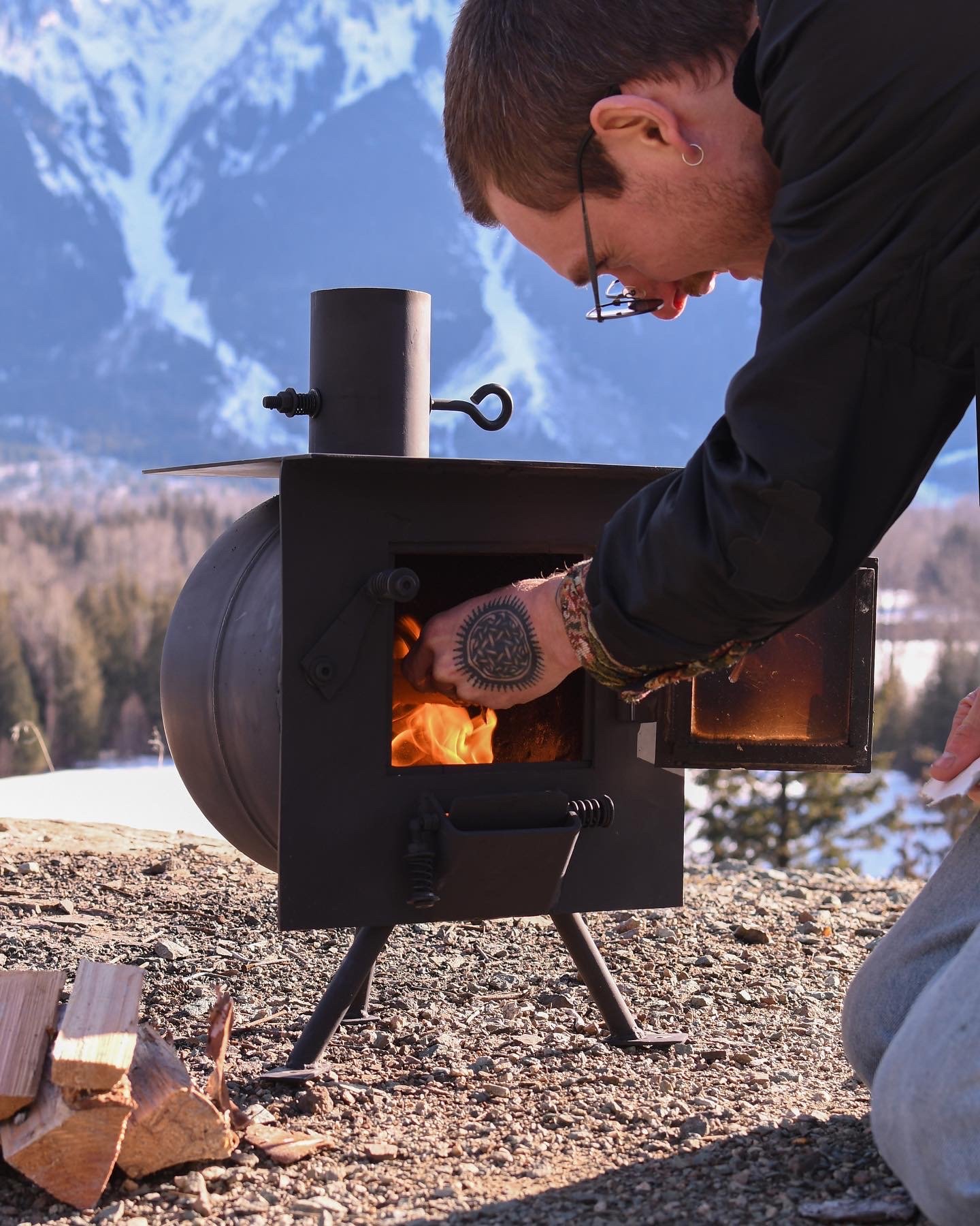 Person tending a small outdoor black metal stove with a fire inside, mountain landscape in the background, person's hand with a tattoo on wrist