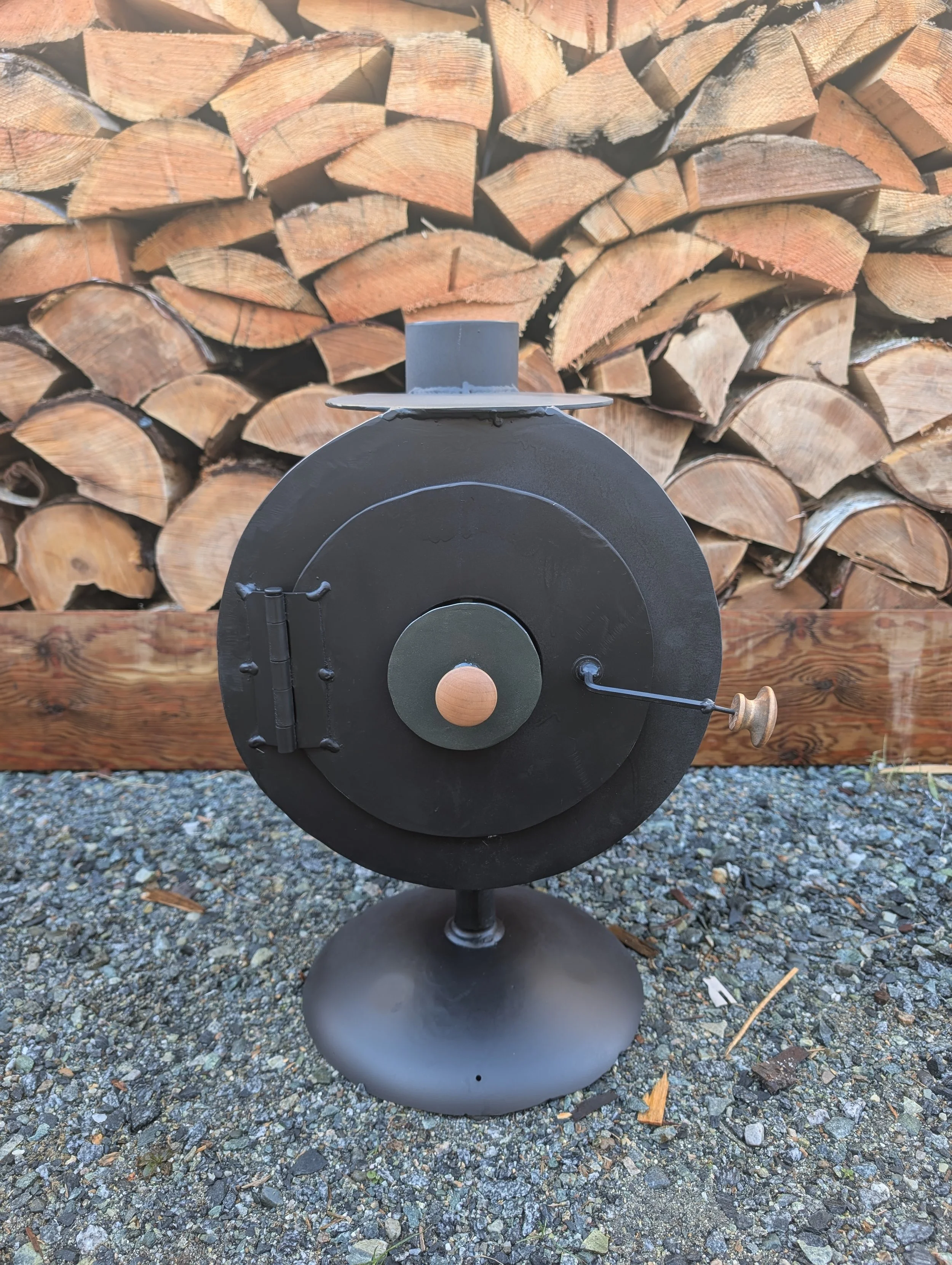 A black metal wood-burning stove on a gravel surface with a wooden log pile in the background.