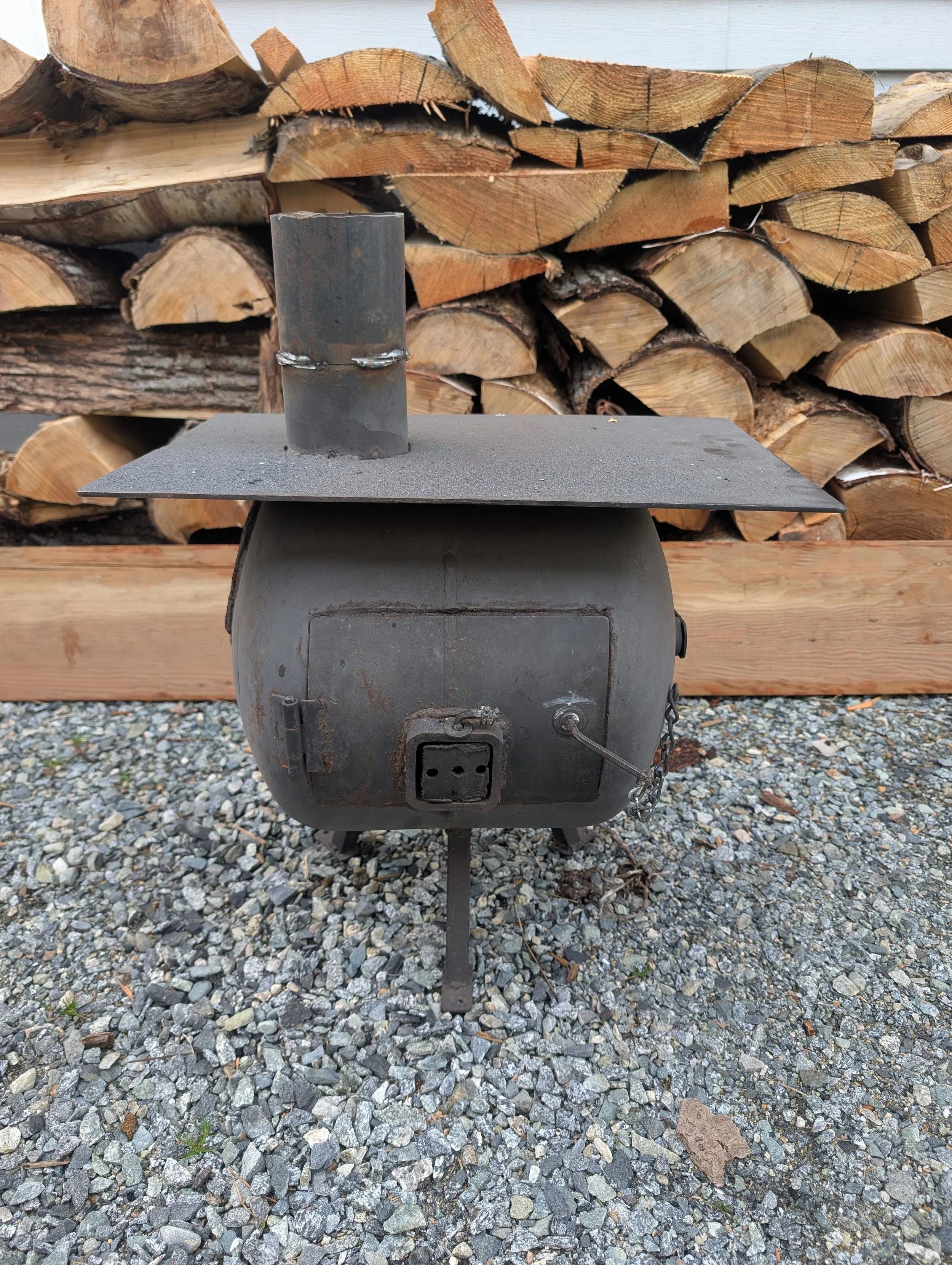 A metal outdoor stove or heater with a chimney pipe and a flat top, placed on gravel in front of stacked firewood logs.