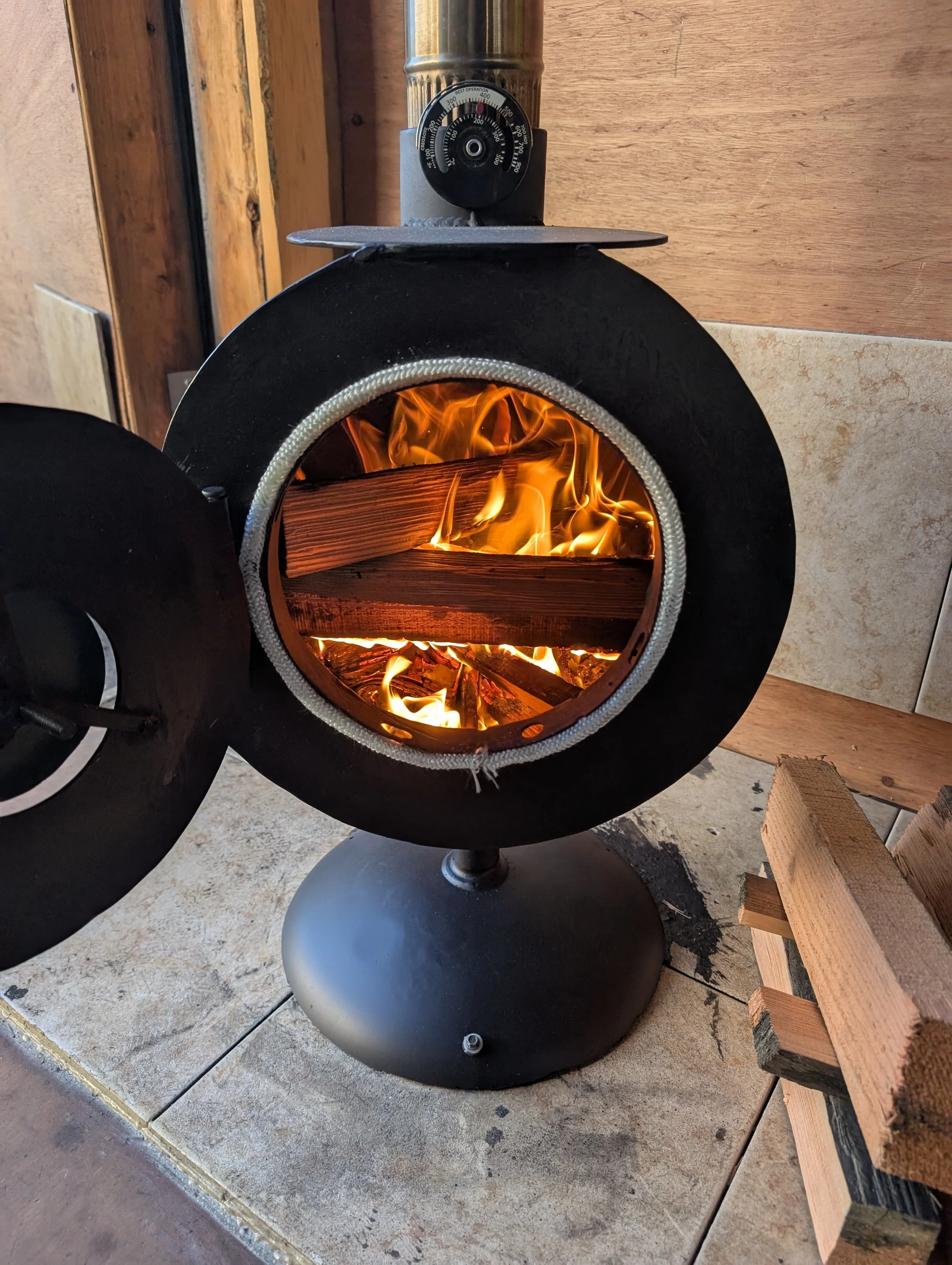 A pellet stove with flames inside, a circular control dial on top, and a metal base. Some firewood is stacked nearby on a tile and concrete floor.