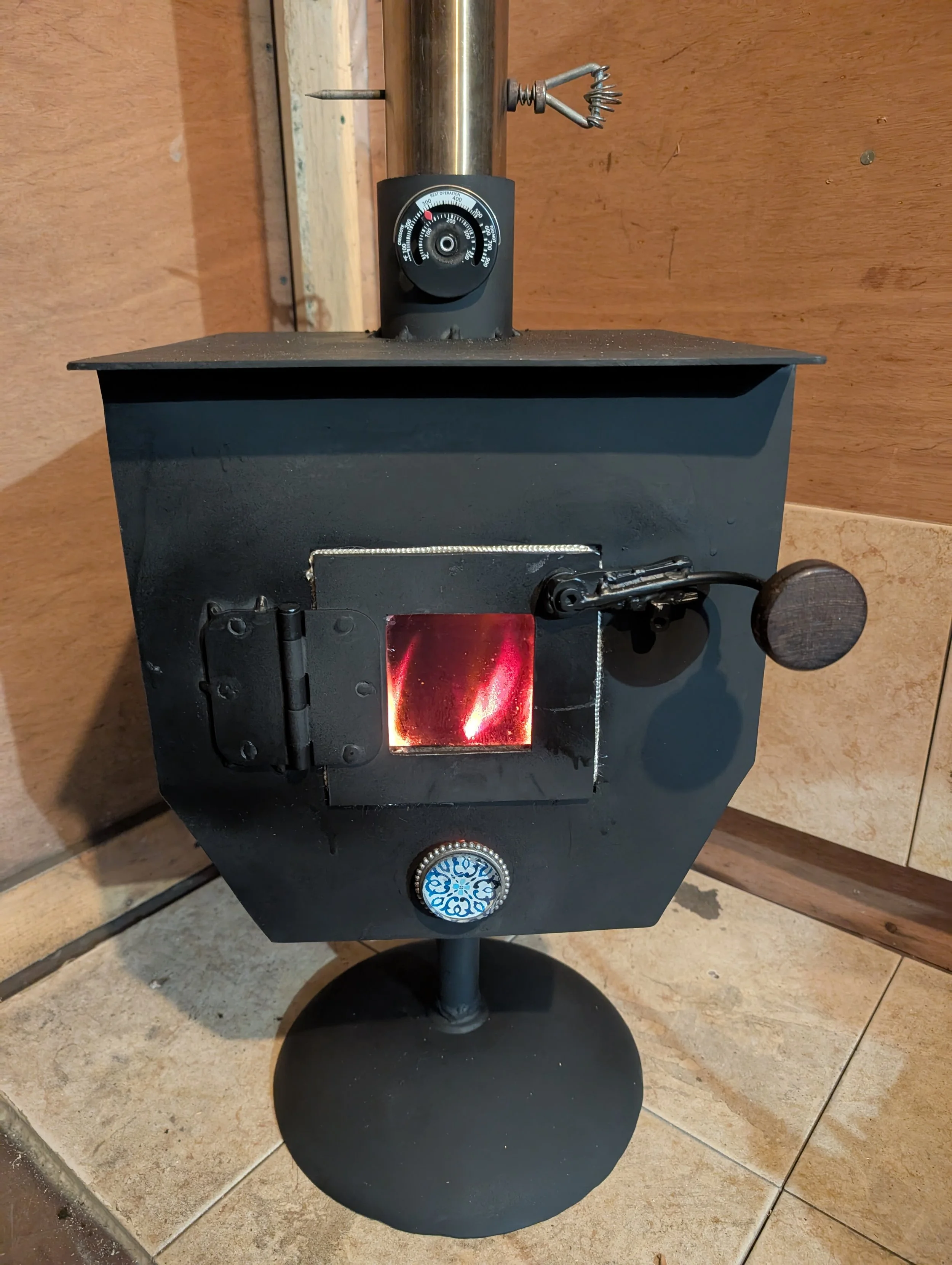 A small black tabletop smoker with a round chimney, a temperature gauge on the side, and a door at the front with an inner red glow indicating hot coals or a heat source inside. It is standing on a stone tiled surface with a wooden wall background.