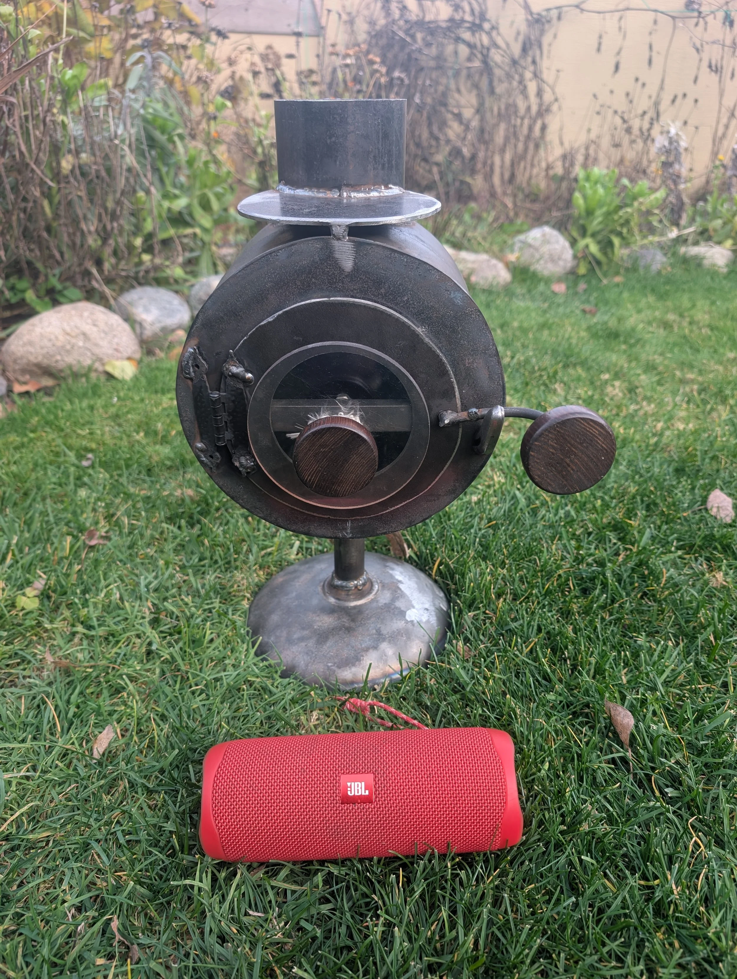 A close-up of a watchmaker's lathe outdoors on grass with rocks and shrubs in background. A red JBL portable speaker is at the bottom of the image.