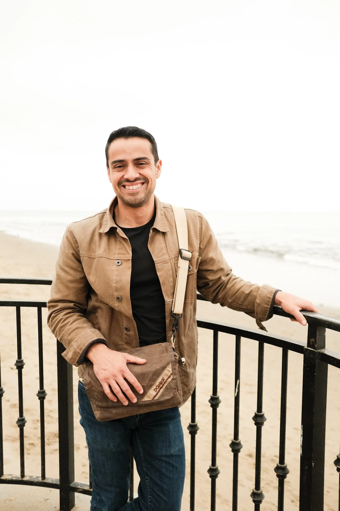 A man with dark hair, smiling, wearing a brown jacket over a black shirt, standing by a black metal fence on a beach with the ocean and cloudy sky in the background.