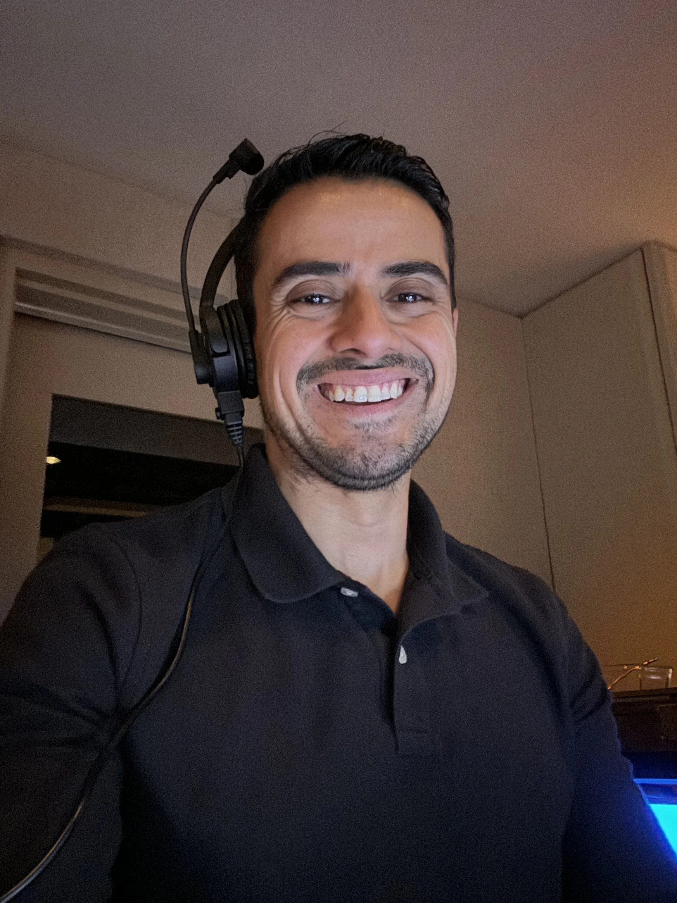 A man with dark hair and a goatee wearing a black polo shirt, smiling, with a headset on, in an indoor setting with warm lighting.
