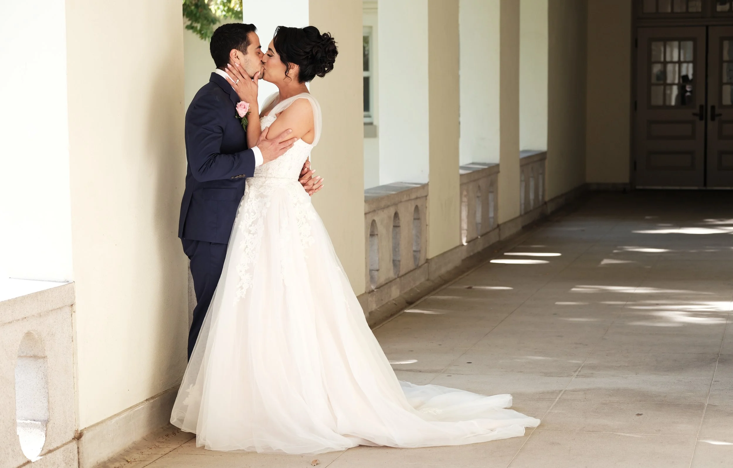 A newlywed couple sharing a kiss, with the bride in a wedding dress and the groom in a suit, standing against a light-colored wall in a corridor with sunlight pattern on the floor.