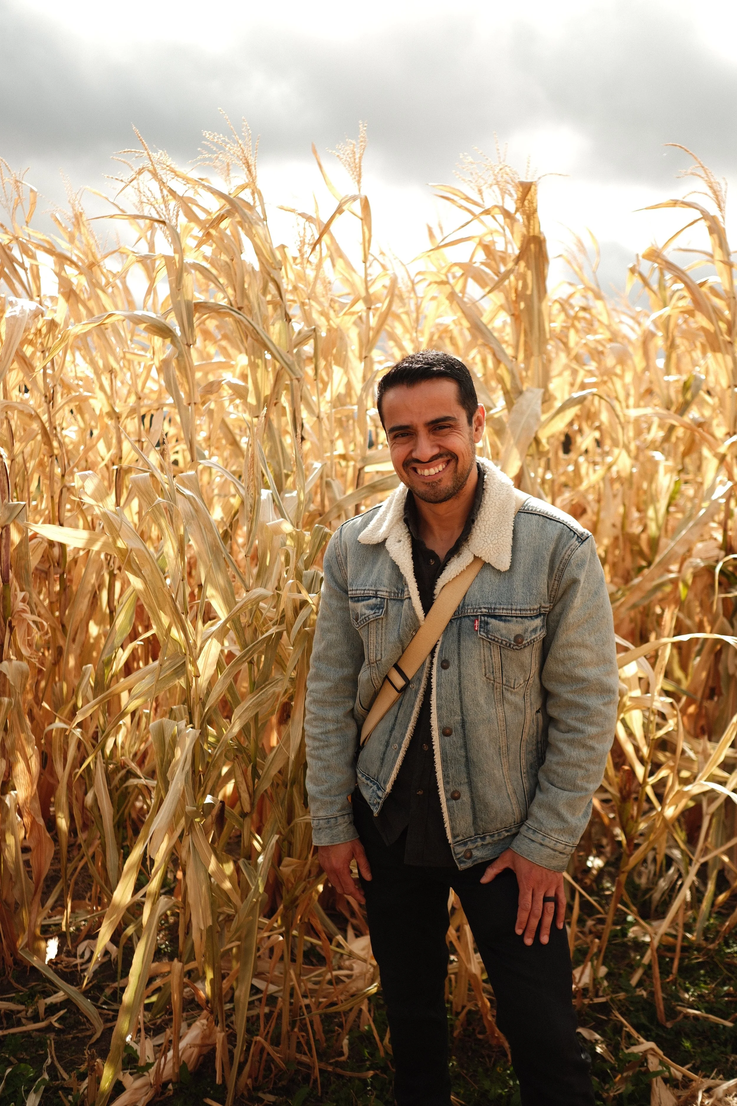 A smiling man standing in a field of dried corn stalks on a cloudy day.