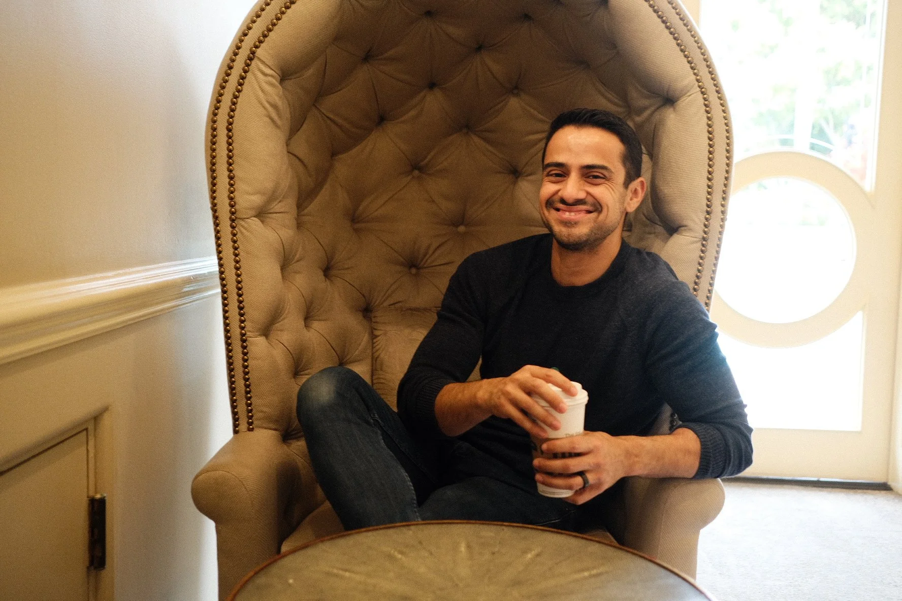 A man sitting in a beige, high-backed, tufted chair holding a paper cup, smiling, with a bright window behind him.
