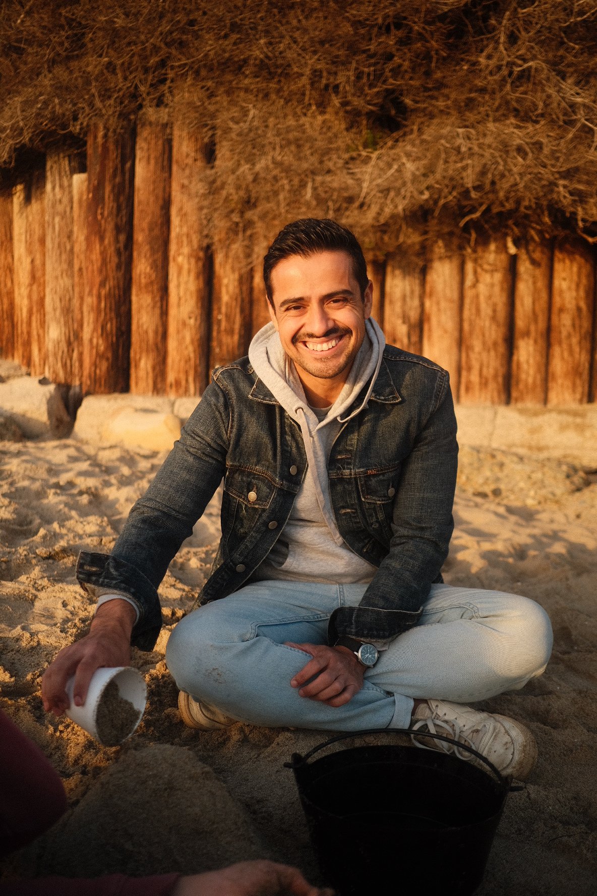 A man sitting cross-legged on the sand at sunset, smiling at the camera, with a wooden fence and dried bushes behind him, holding a cup of sand.