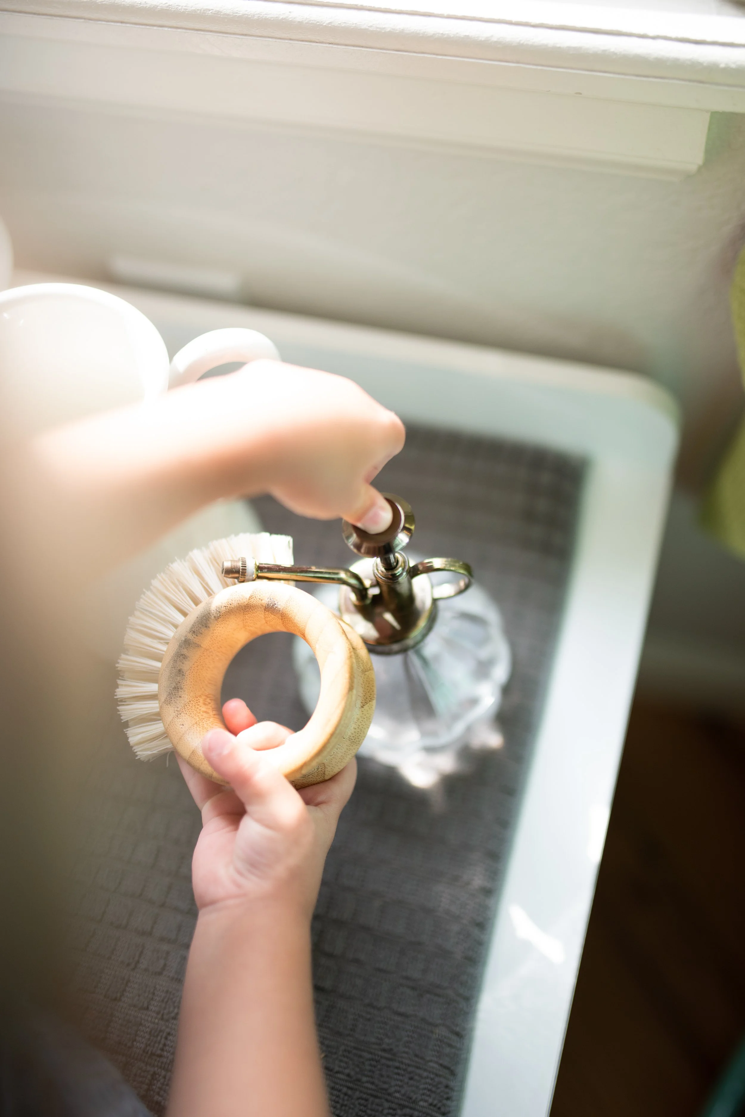 A person holding a wooden ring with a scrub brush attached, near a sink with a faucet and a glass soap dispenser, in a kitchen or bathroom setting.