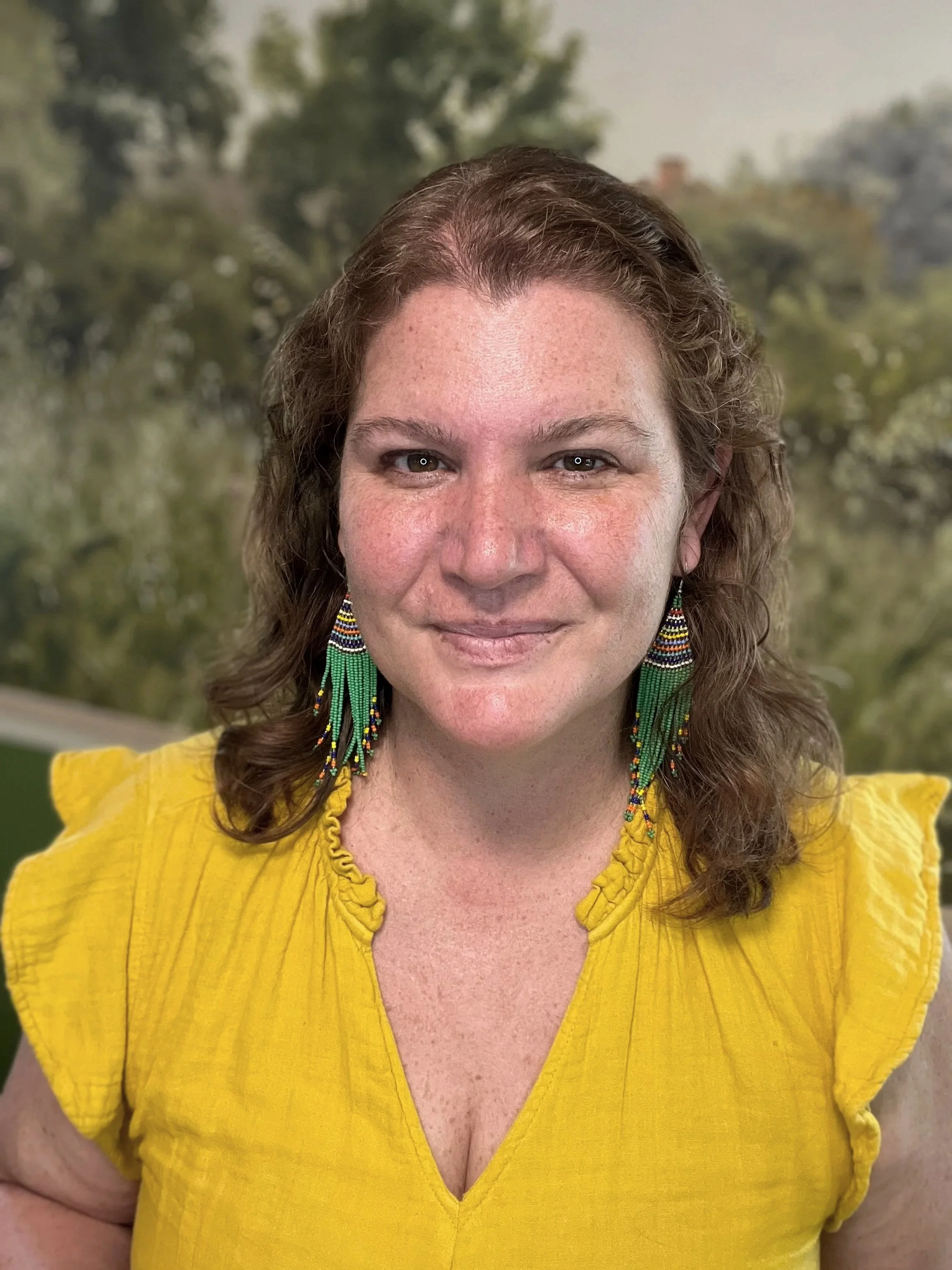 A woman wearing a yellow top with ruffled sleeves and multicolored beaded earrings, smiling, with a blurred outdoor background.