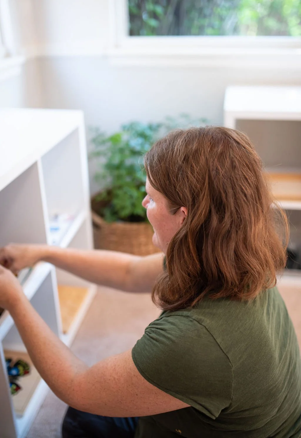 A woman with red hair wearing a green shirt, reaching into a white shelf inside a bright room with a window and green plants in the background.