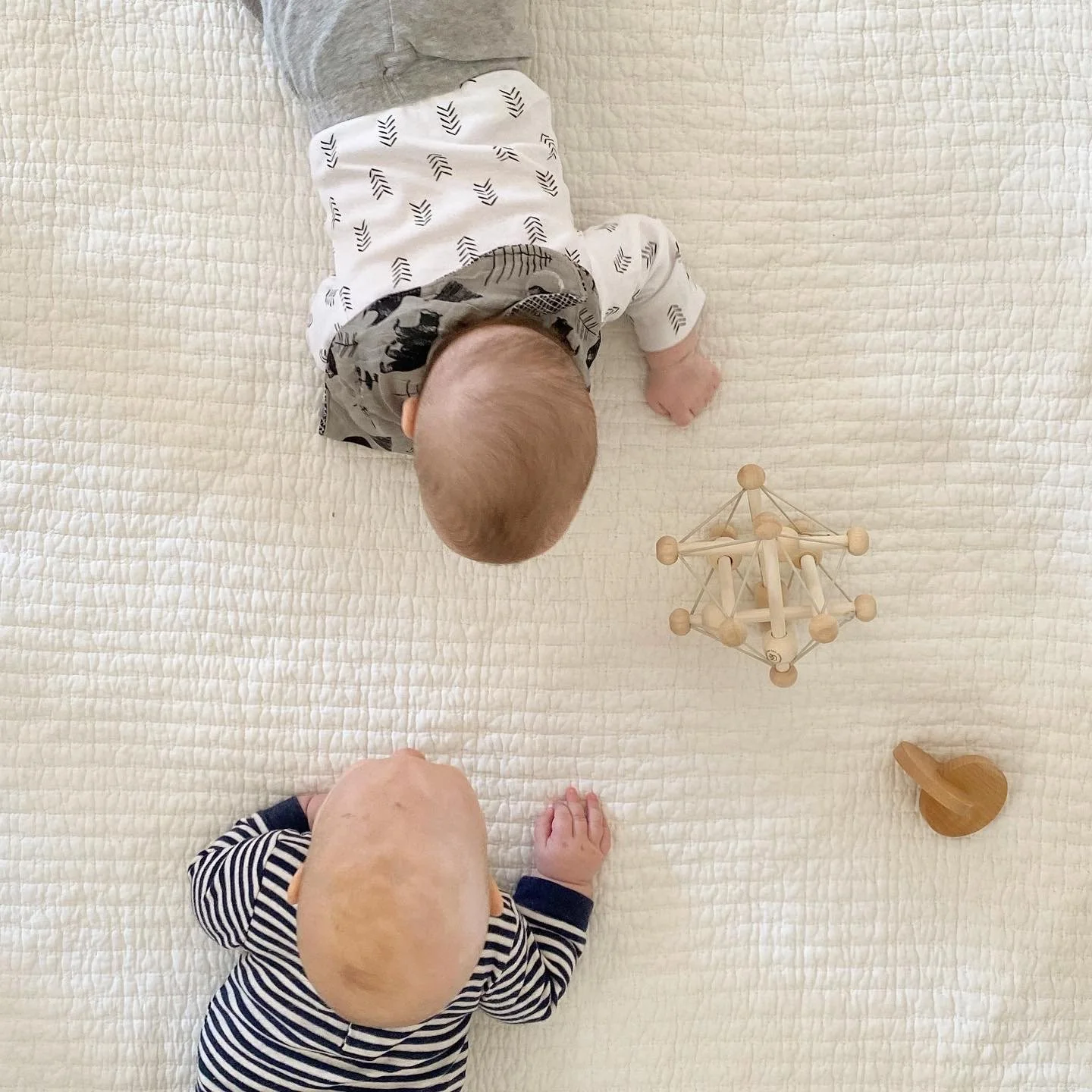 Two babies crawling on a white quilted mat, playing with wooden toys.
