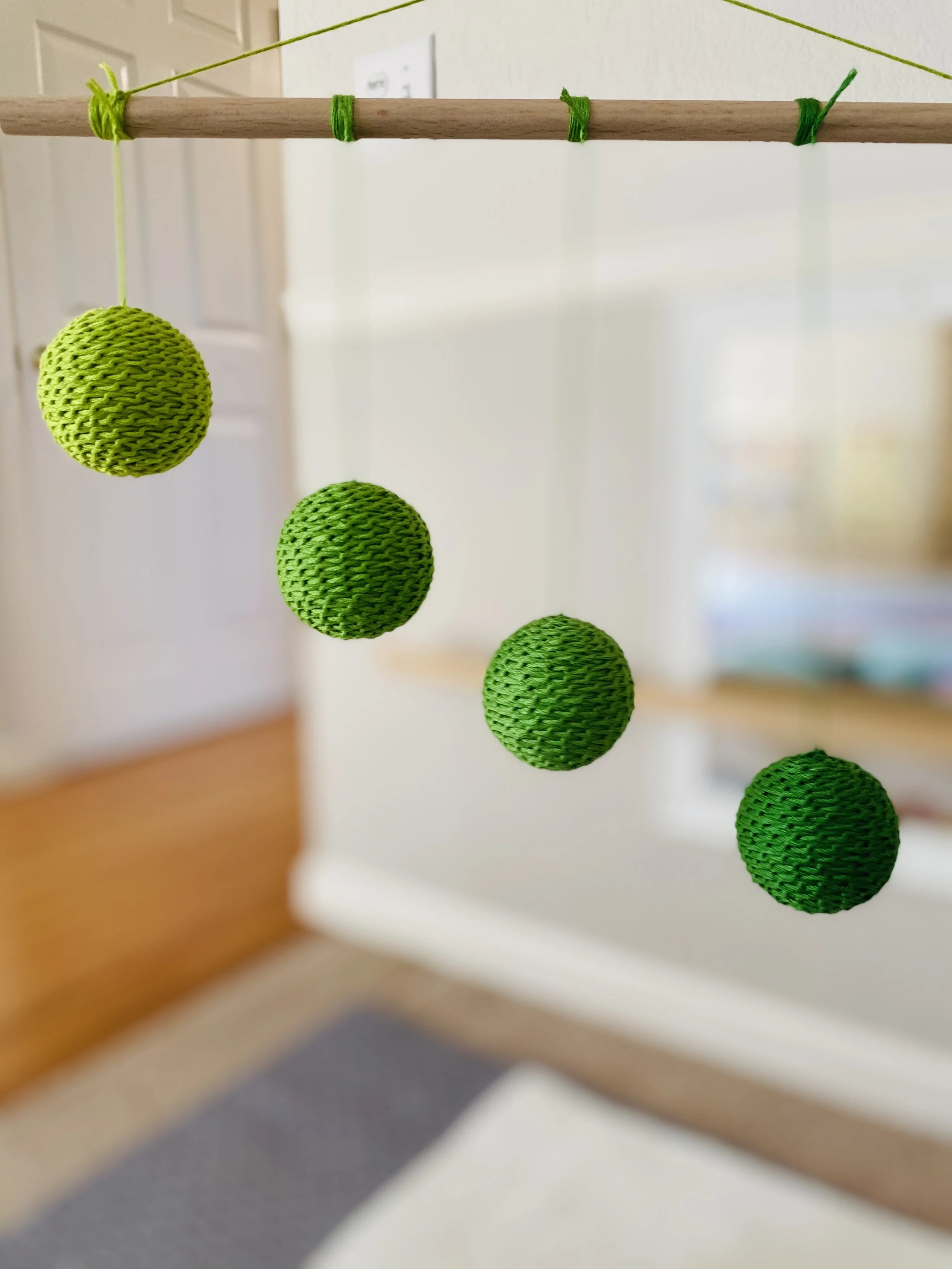 Close-up of a hanging mobile with green textured balls and green string, attached to a wood stick, indoors with part of a room visible in the background.
