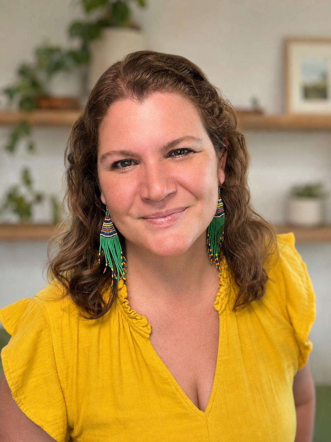 A woman with curly brown hair and blue eyes, smiling, wearing a yellow top and colorful beaded earrings, in a room with plants and framed pictures in the background.