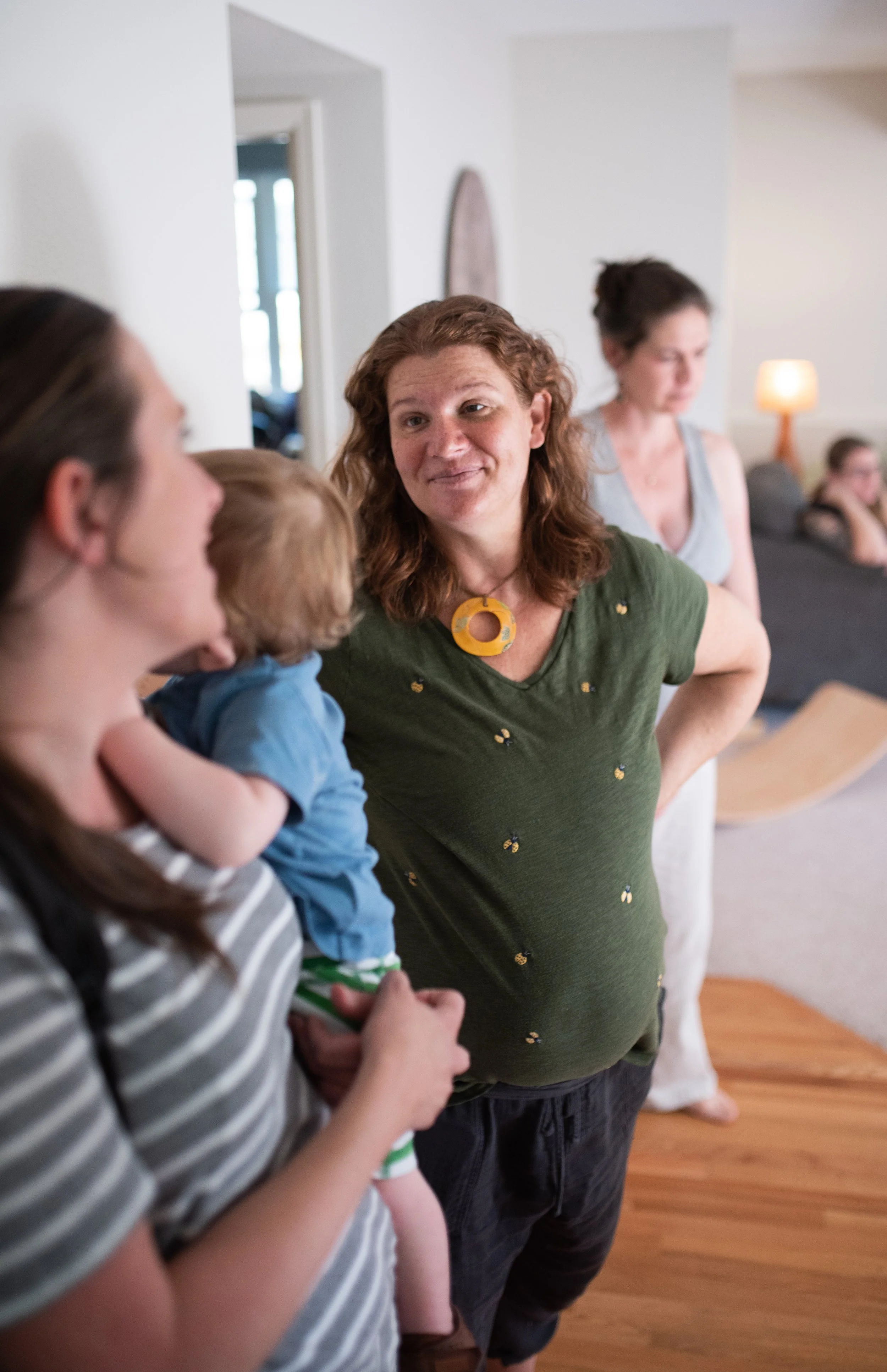 A woman with curly red hair wearing a green shirt with small yellow patterns is engaged in conversation with a woman holding a young child. The background features a living room with a gray sofa, a warm lamp, and a few other people sitting.