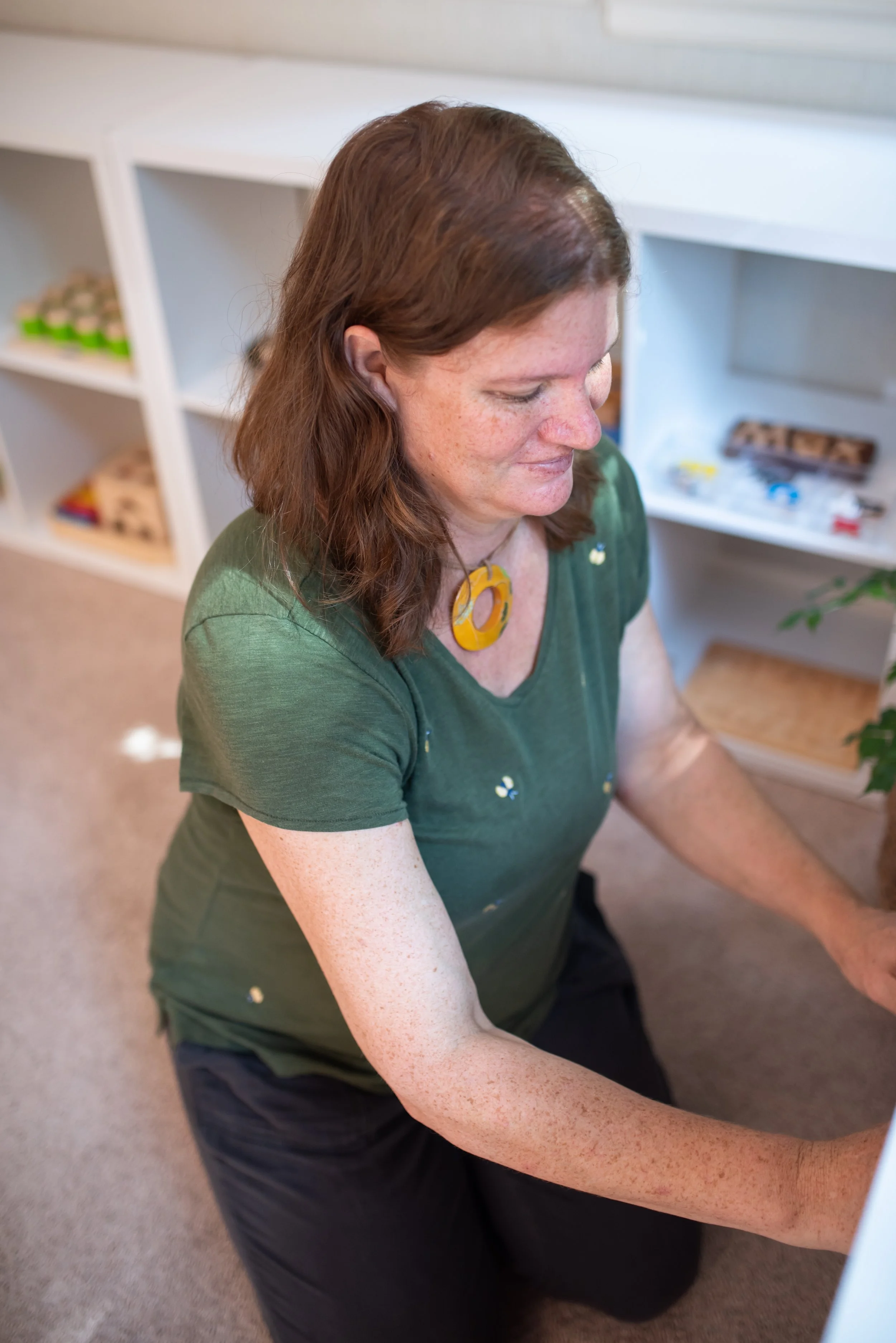 A woman with shoulder-length brown hair, freckles, wearing a green shirt and a yellow circular necklace, is kneeling on the floor in a room with white shelving units in the background.