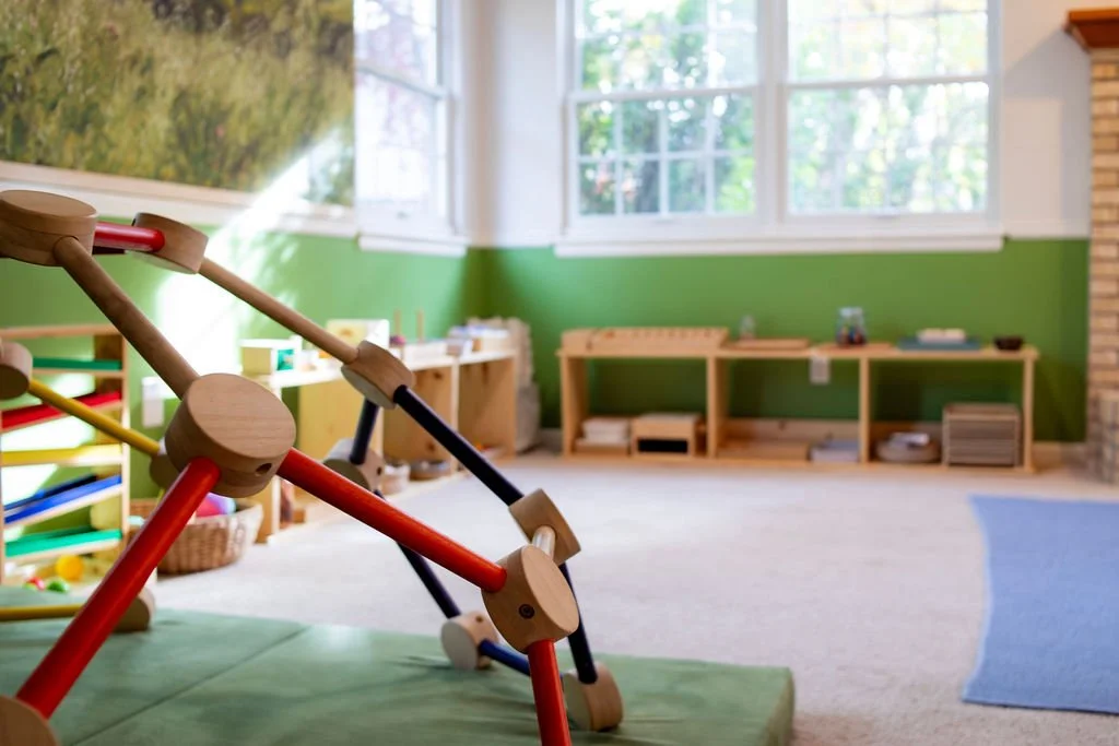 Indoor playroom with climbing structure, wooden toy shelves, and large window with outside view.
